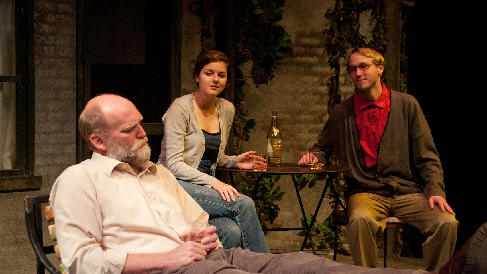 An old man sits in a chair looking low and outward toward stage left. While a younger man and woman sit behind him at a table, looking at the older man. The woman has a look of soft amazement on her face while the young man smiles. 