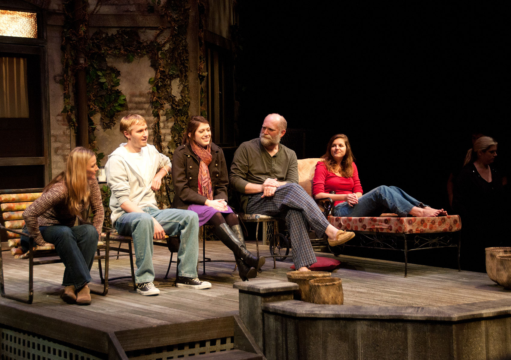 Five characters, three women and two men, sit in four porch chairs and a lounge chair in a line on stage. They all face toward the middle as if they are having a conversation with each other.