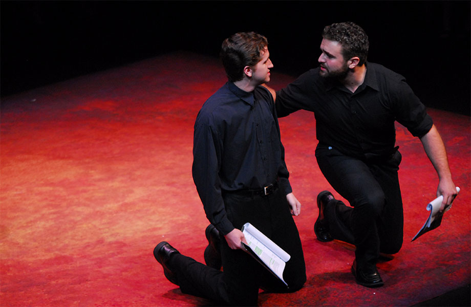 Two men dressed in black are kneeling on the ground, one has his hand on the other’s shoulder and is talking to him and each man is holding a script.