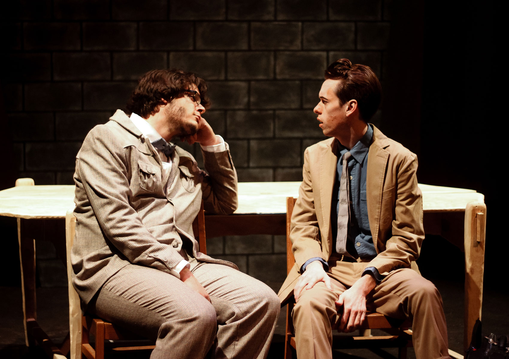 Two men are wearing suits and sitting in chairs, one man is staring at the other as if in a reverie while the other is looking back at the first with concern, mouth slightly ajar.