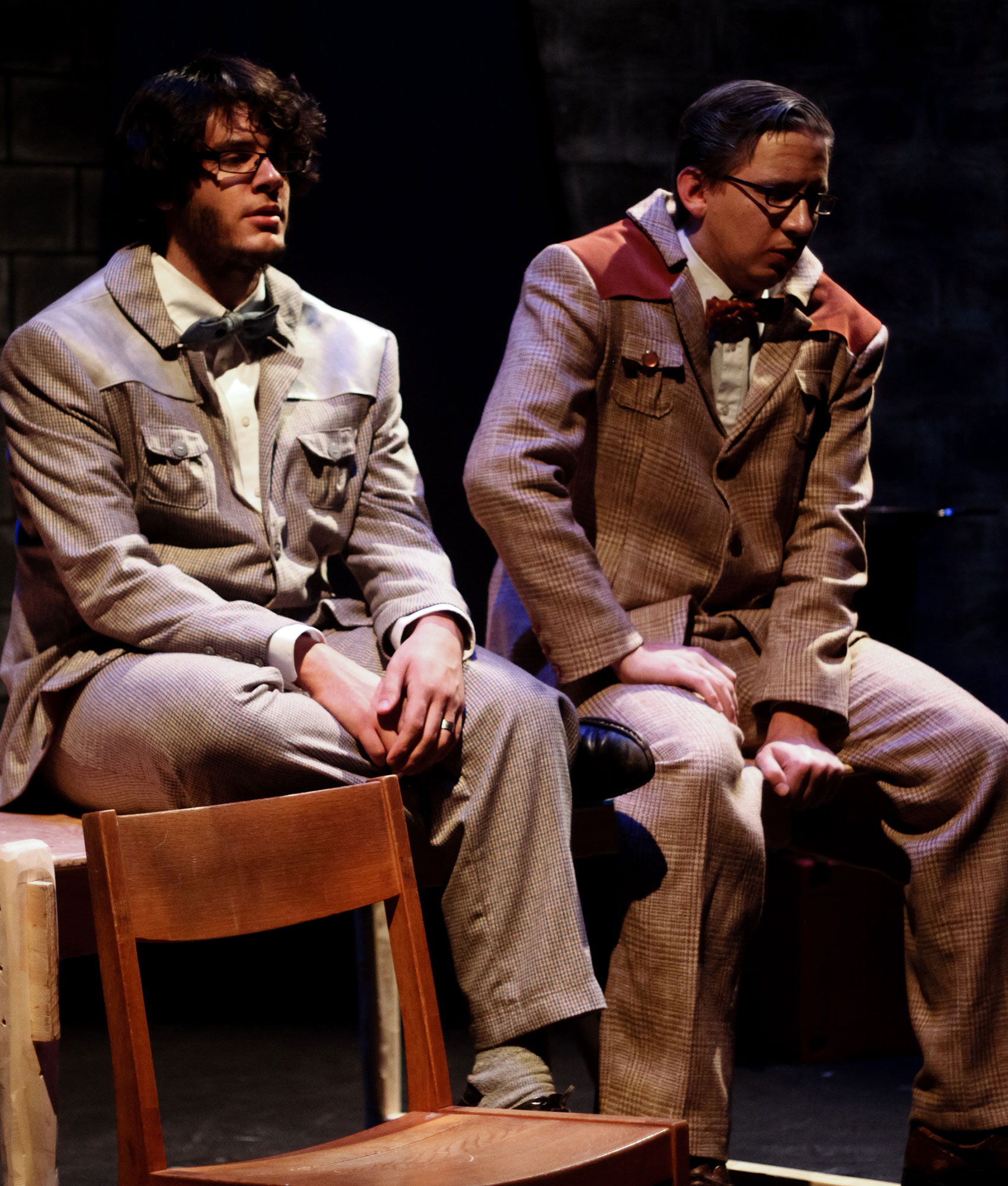 Two men sit next to each other on a table-top. One has a contemplative expression on his face while he has his hands crossed in his lap while the other is hunched forward with a look of worry on his face, his right arm bent slightly with that hand on the right knee while his left arm is straight and that hand clutching the edge of the table tightly.  