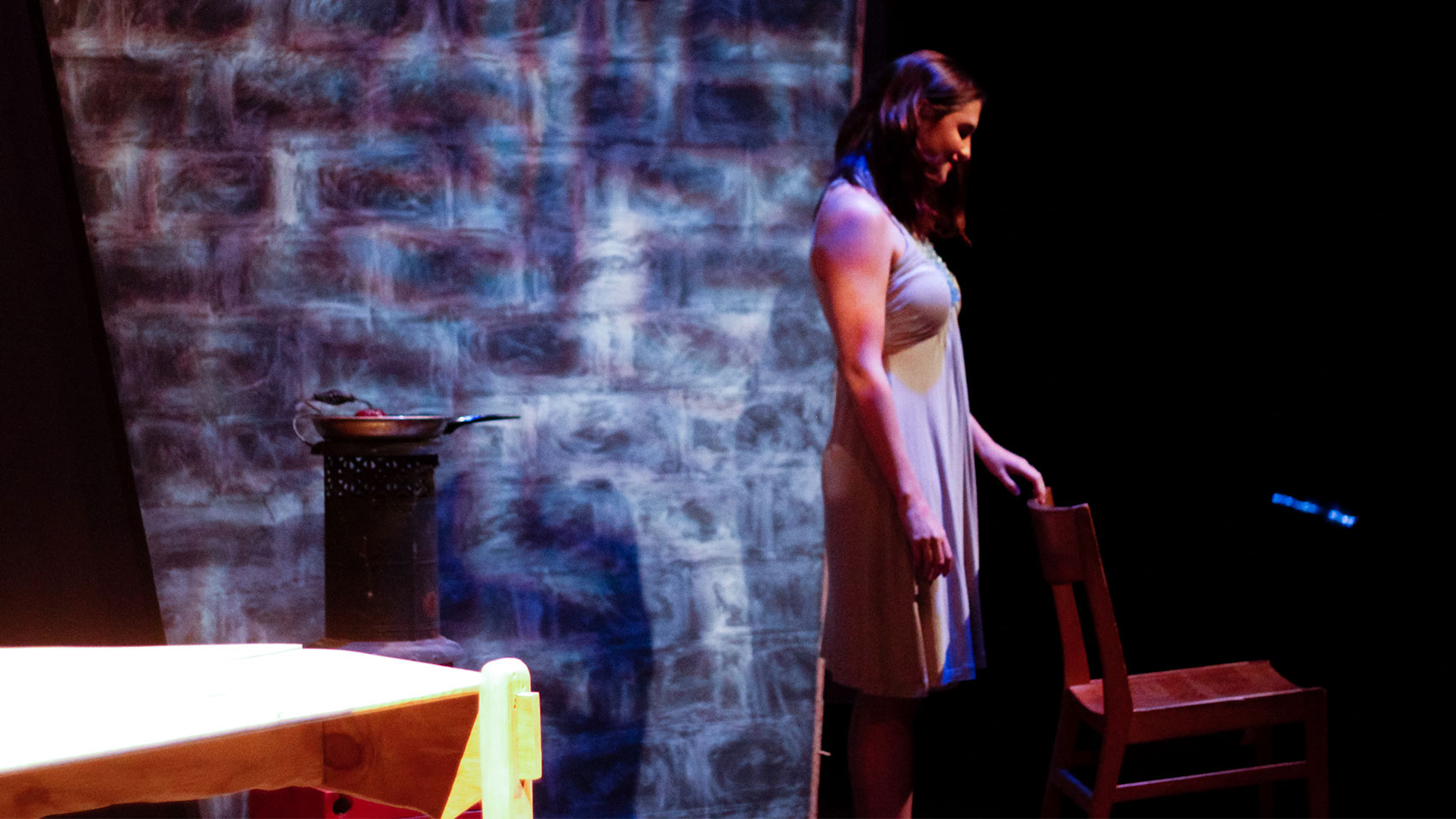 A woman smiles while looking down toward a wooden chair, letting her fingertips brush the back of it gently.