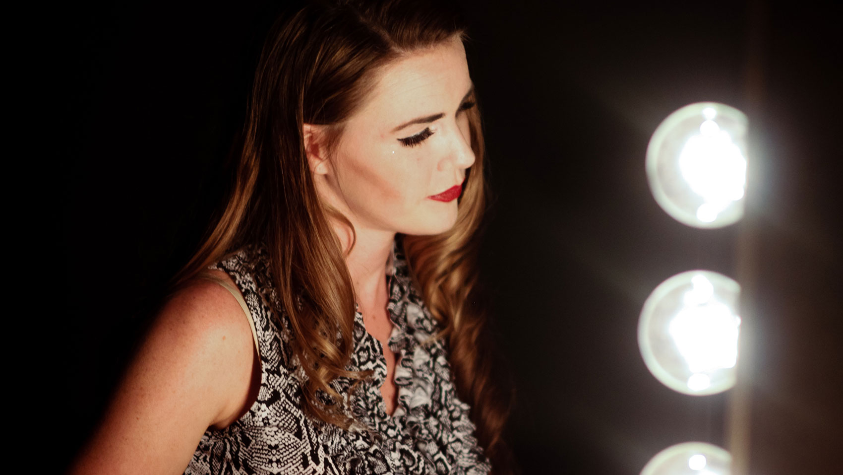 A woman stares into a dressing room mirror lit with large round bulbs.