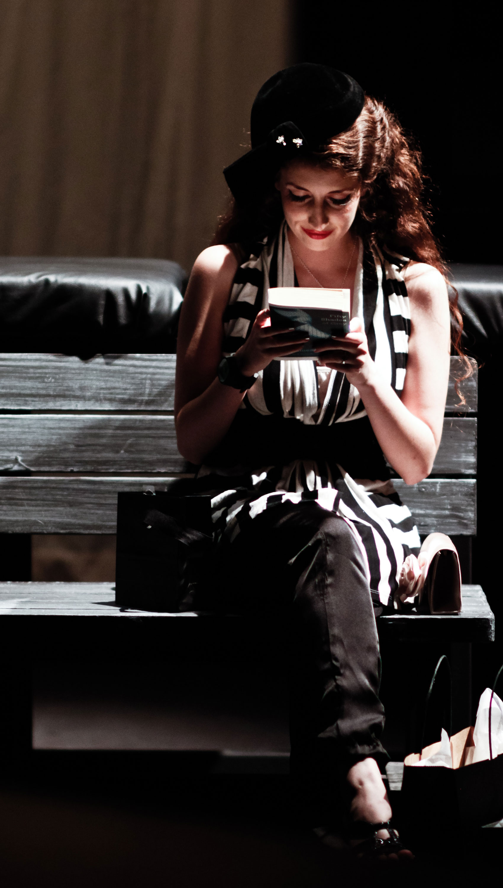 A woman is sitting on a wooden bench with a bag on one side of her and a pocketbook on the other. She is smiling somewhat and is reading the back of E.L. James’ Fifty Shades of Grey.