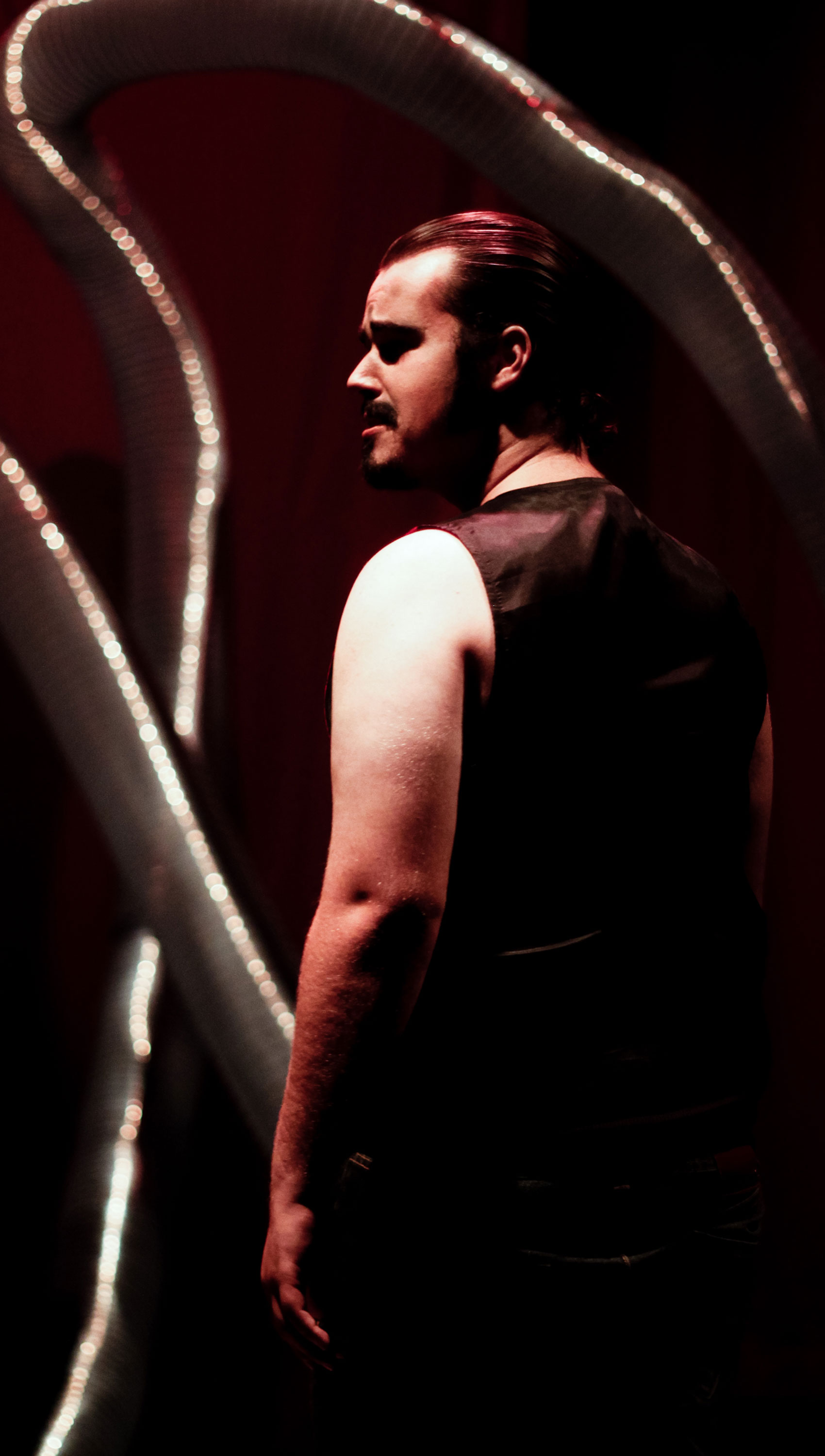 A man standing in front of a thin strip of looped LED lights in the background is facing stage right. Part of him is illuminated and part of him is silhouetted; he is wearing all black clothes.