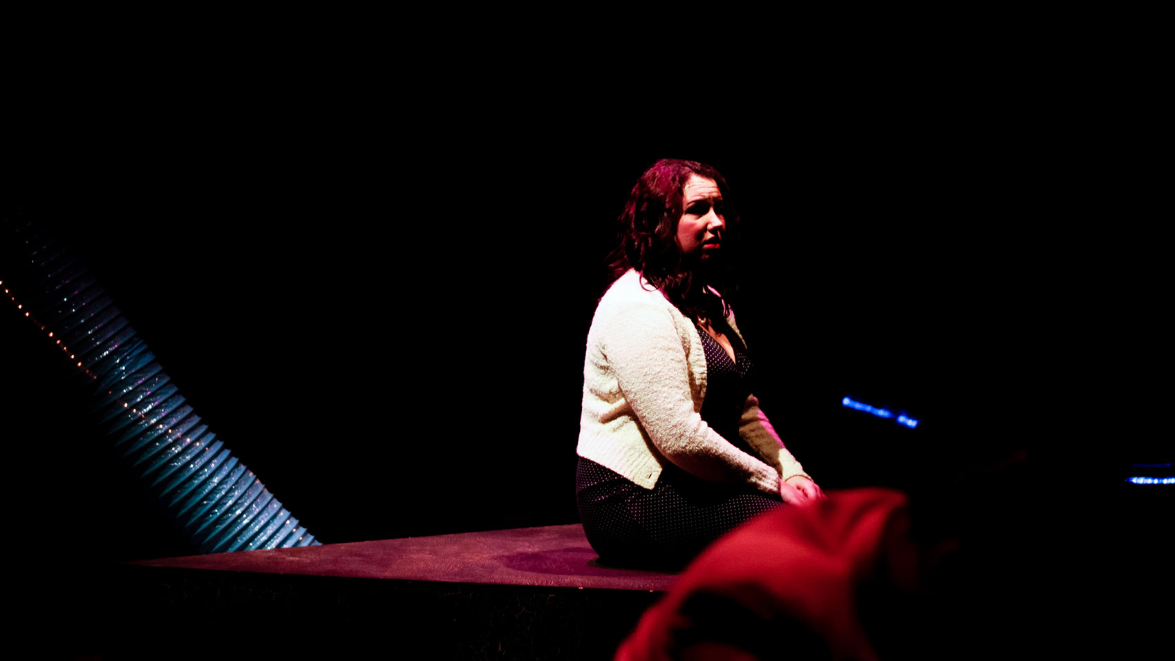 A woman sits upright with her hands crossed in her lap and a hurt look on her face as she sits on a carpet-covered platform. 