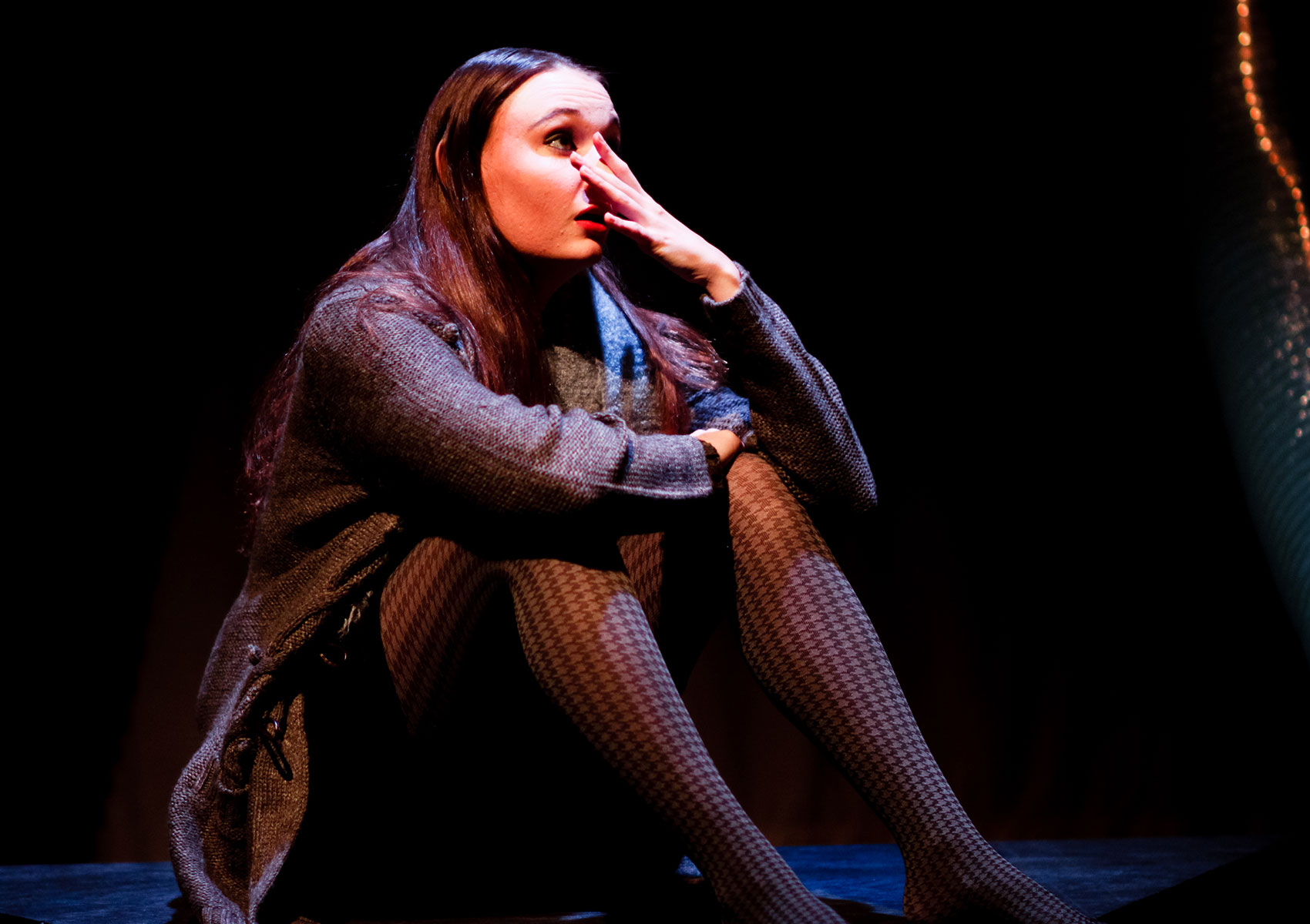 A woman sits on a platform with her knees to her chest. Her right arm and hand are resting on the opposite knee while she holds her left hand to her face. She is looking out over her fingertips toward stage left.