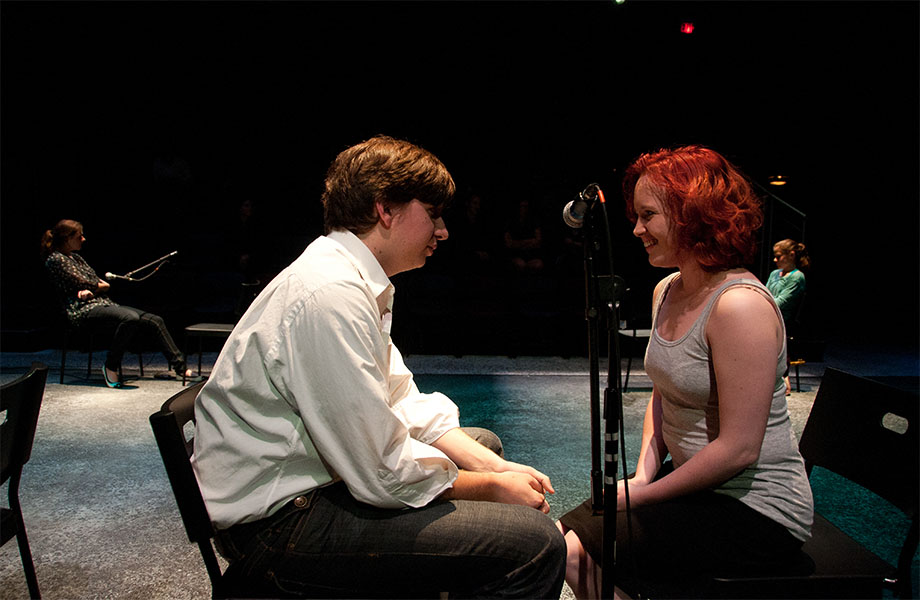 A male and a female character are smiling at each other with a microphone stand in between them. There are two characters in the background, another one is next to a microphone stand.