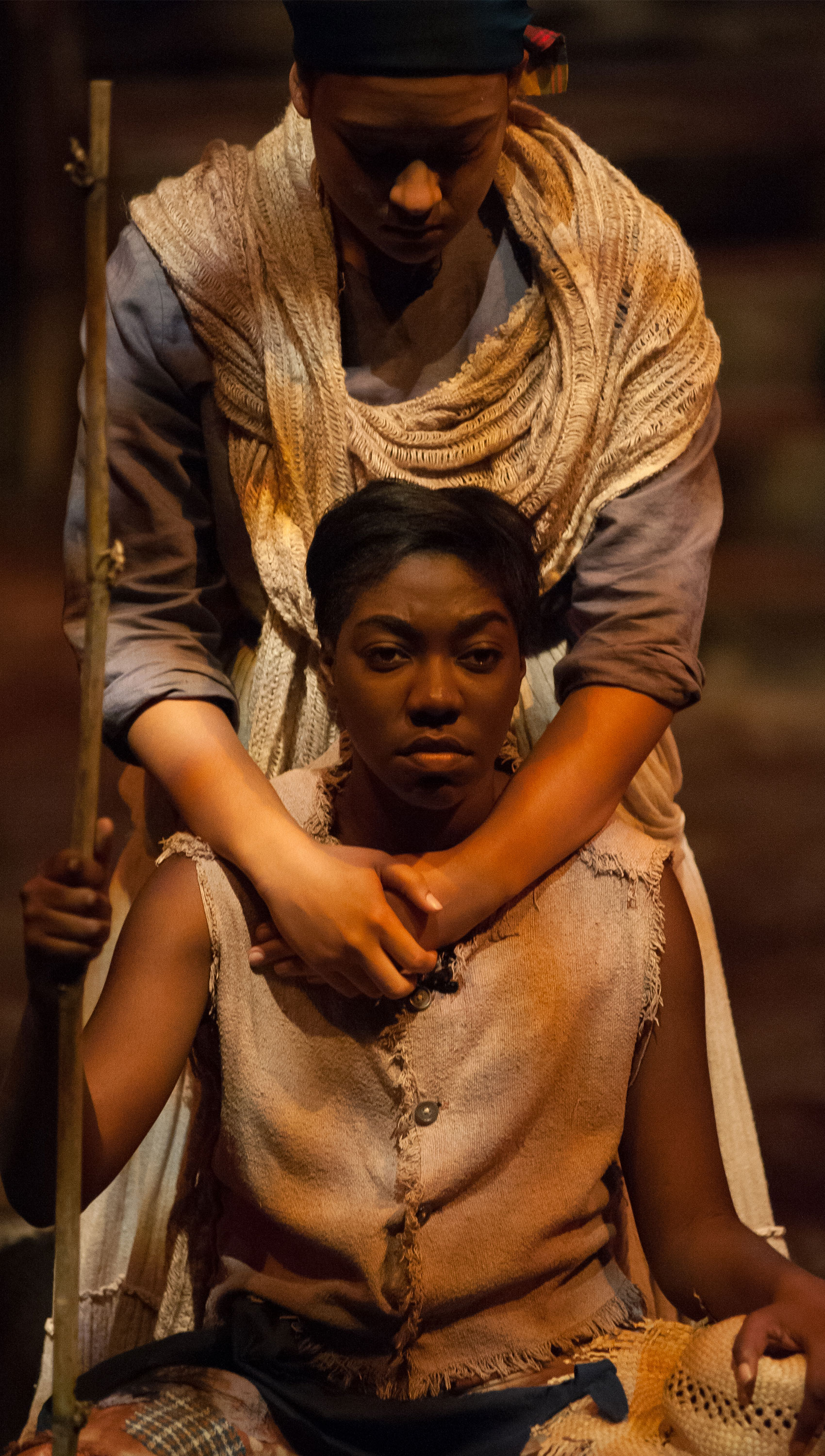 A woman sits, holding a stick erect in her hand, facing the audience. Another woman stands over the first woman with her hands clasped over the first woman’s chest. 