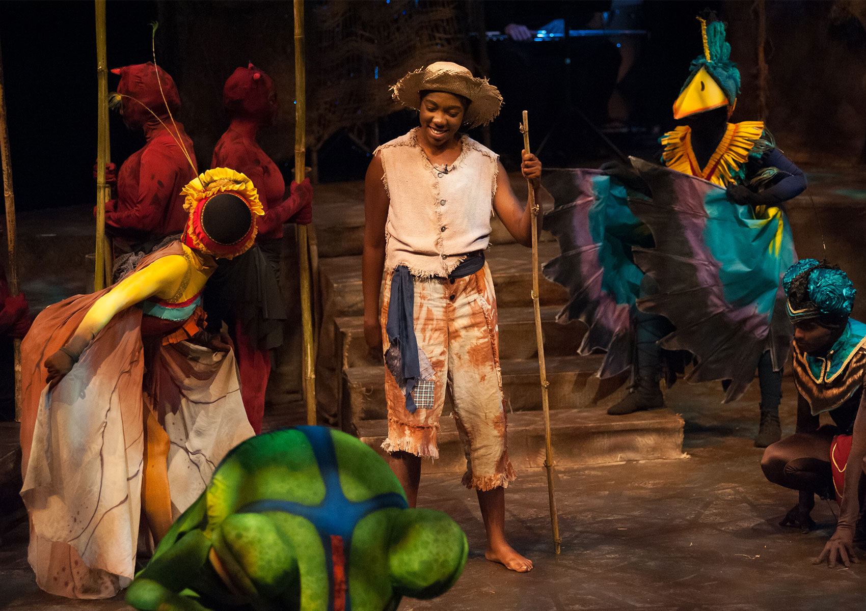 A man dressed as a farmer smiles at characters dressed as a bee and a frog. A character dressed as a bird and another insect watches this exchange while two other characters in red costumes stand back to back behind the farmer holding poles erect. 