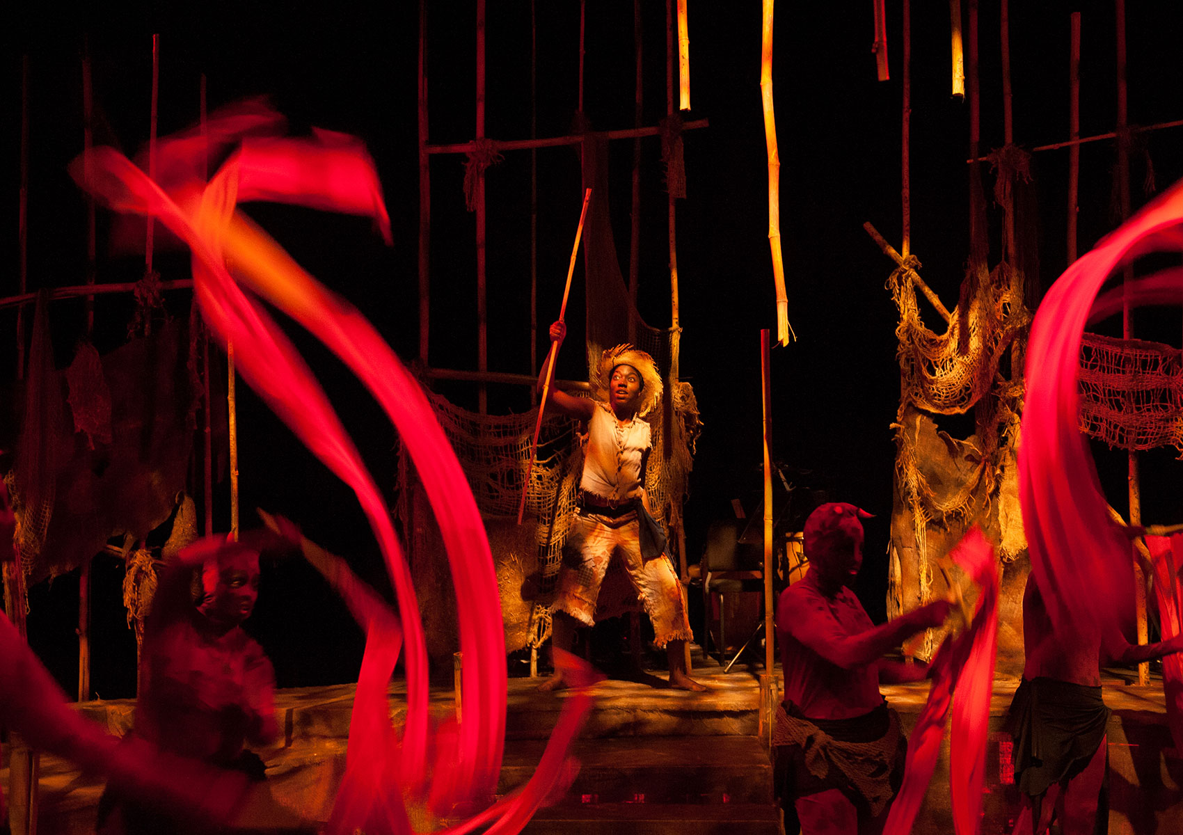 A man dressed as a farmer holds a pole in fear as two characters in red costuming wave red silk ribbons in the air.