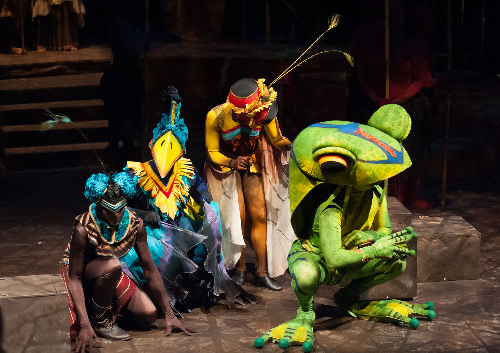 A group of four characters are grouped onstage, one dressed as a bird, one as a frog, and the other two are dressed as creatures that resemble insects or birds of some kind by wearing vibrant colors and feathery antennae appendages.