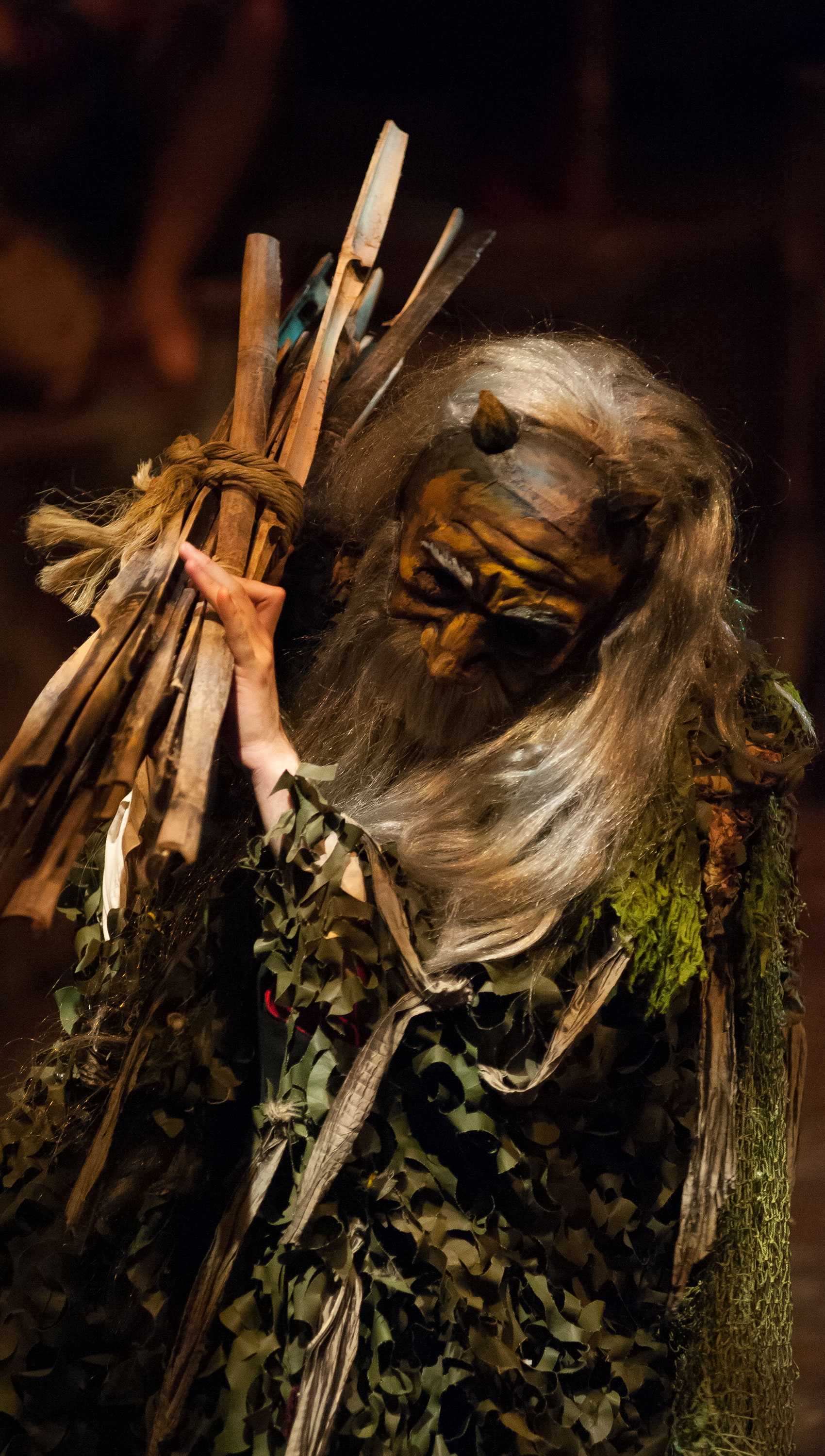 A character with a mask carries a bundle of sticks on their shoulder.