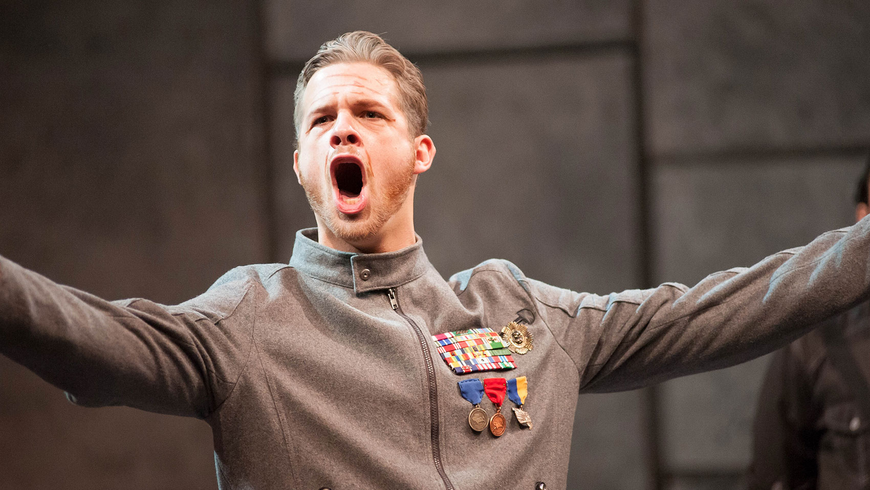 A man is projecting with his arms outstretched. He is wearing a grey coat covered in medals.