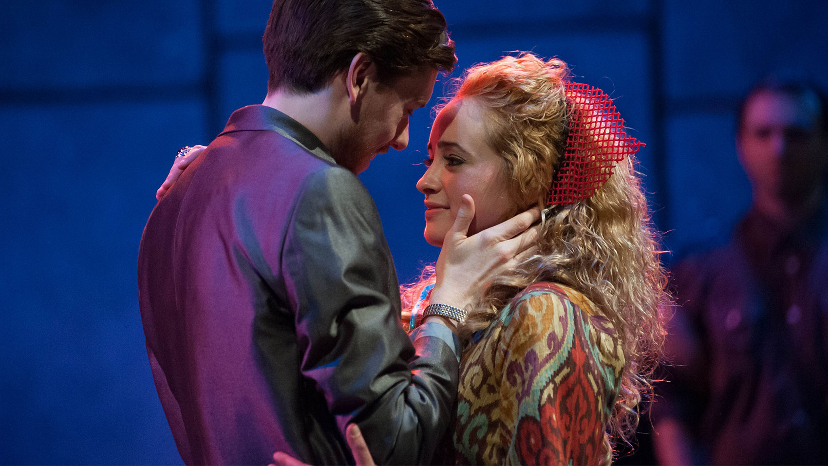 A man and woman with a red hair accessory are holding each other wearing vibrantly colored costumes. He is looking lovingly at her while one hand can be seen caressing her cheek with his thumb. She is looking at something behind him, and is holding the man with one arm on his right shoulder and the other near his waist.