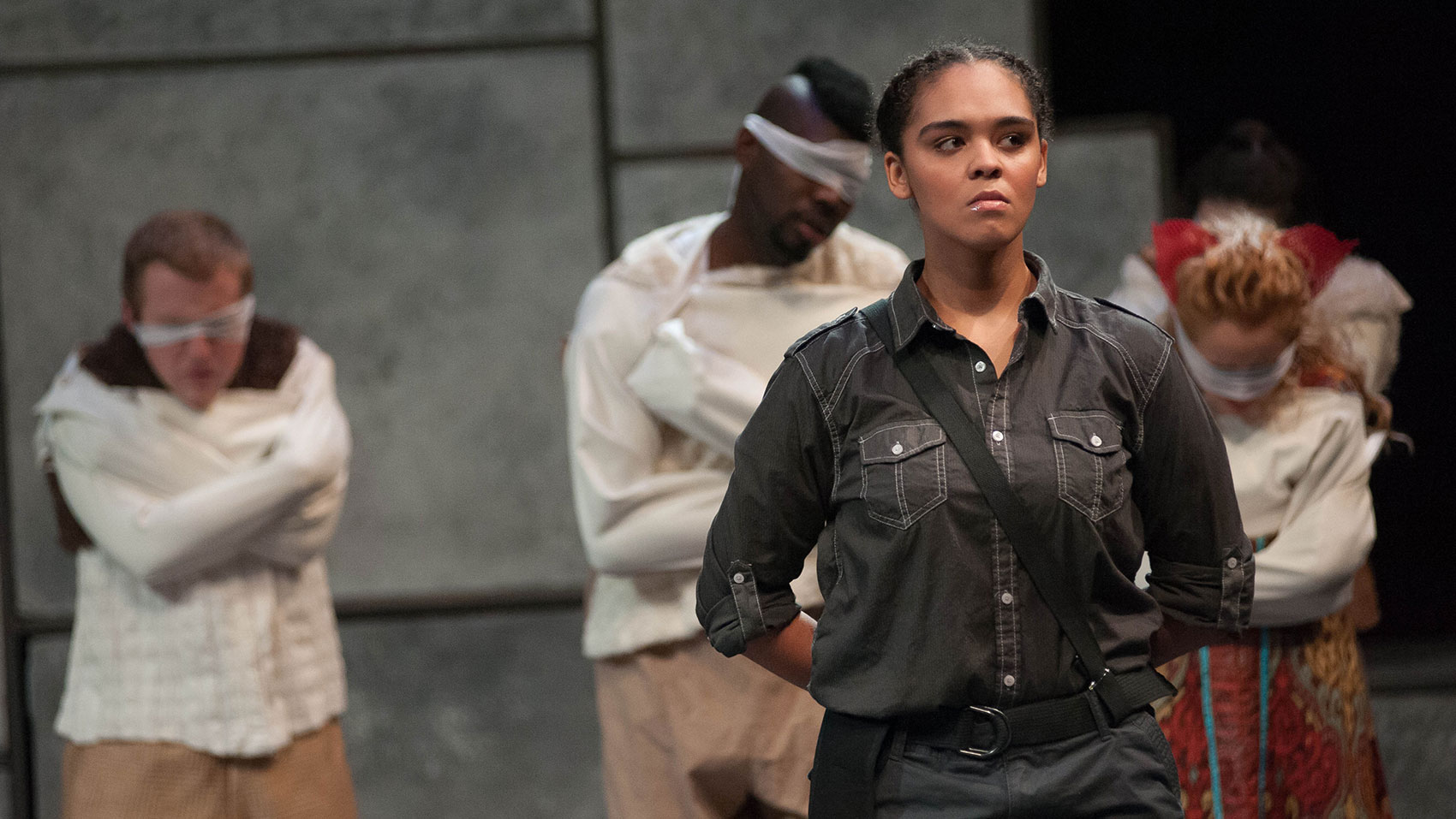 A woman in dark grey clothes stands at attention with her hands behind her back and a stern look on her face. She stands in front of three characters, who are blindfolded and bound in straightjackets. Another character, facing away from the audience, is behind one of these three characters.