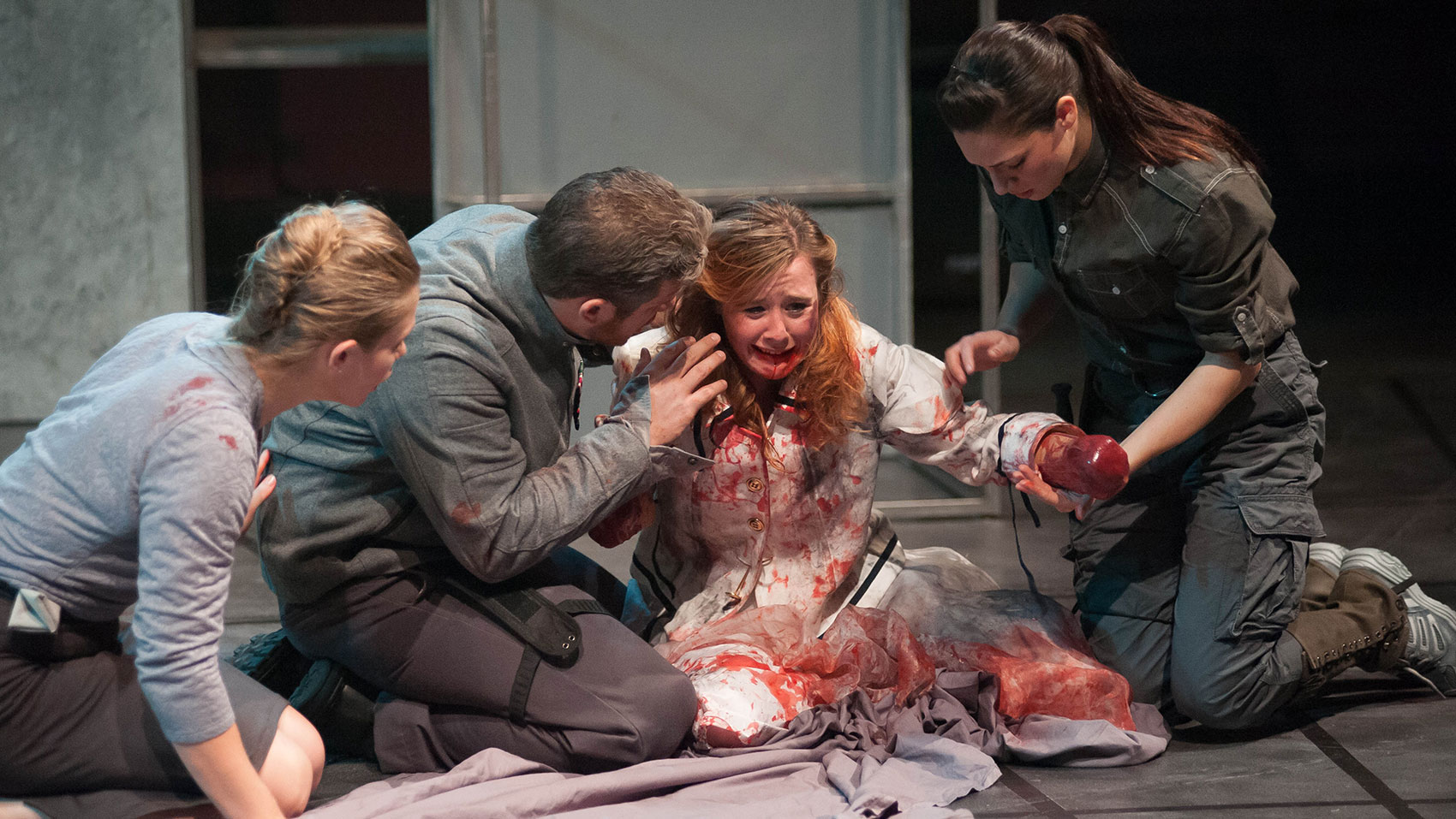 A woman wearing white is covered and blood, crying, with one of her arms ending in a bloody stump, she is kneeling on a sheet and a woman is on her knees next to the one-armed woman. This woman is holding the stump in one hand as she looks down on it and the other arm hovering over that. A man is kneeling on the other side of the one-armed woman and is speaking to her with his hand raised slightly and holding onto her with the other. Another woman is kneeling on the other side of the man, her hand on his side and propping herself up with the other hand; she looks as if she is speaking to someone. 