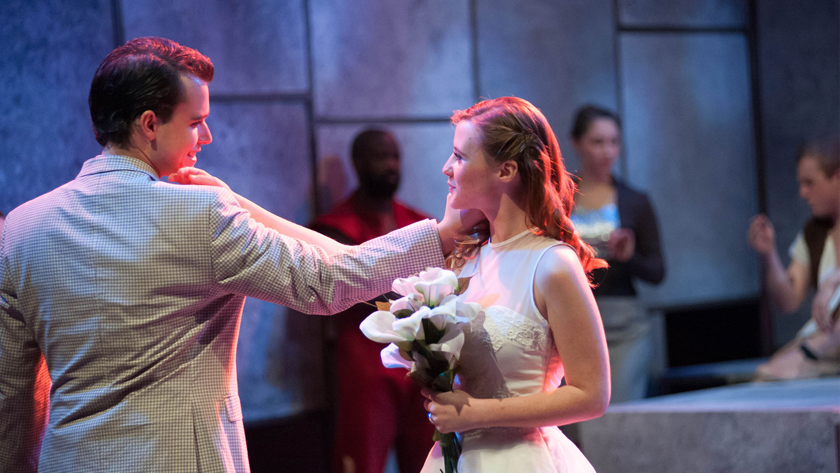 A man and a woman stare at each other lovingly. The man is facing away from the camera but his back indicates he is wearing a suit jacket while staring at the woman from his profile, using his left hand to caress her left cheek facing away from the audience. The woman is wearing a white dress and holding calla lilies in her right hand while caressing the man’s left cheek that faces the audience with the knuckle of her index finger.