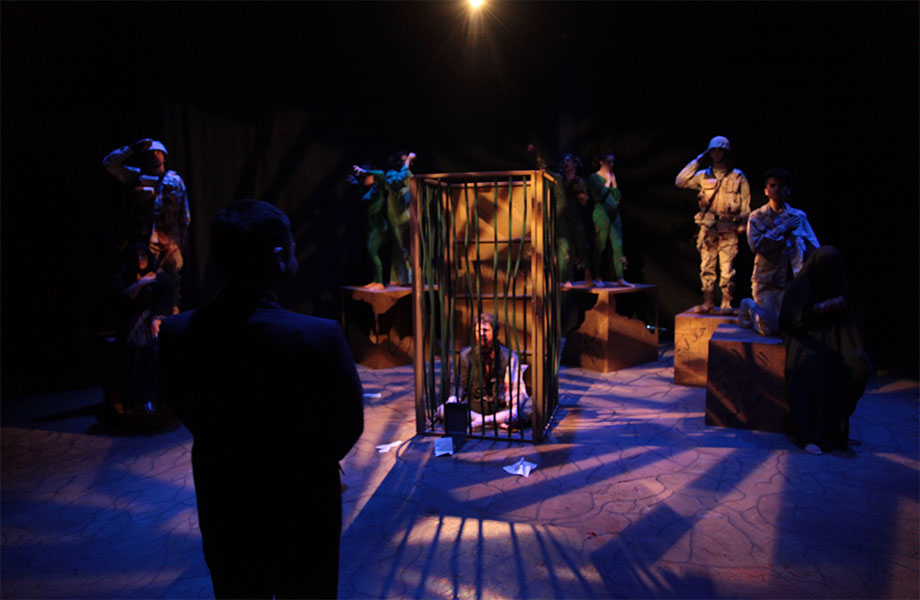 A man is in a cage surrounded by other characters on boxes, some characters are dressed as soldiers while others in the background are wearing solid green. A woman is watching the man in the cage.