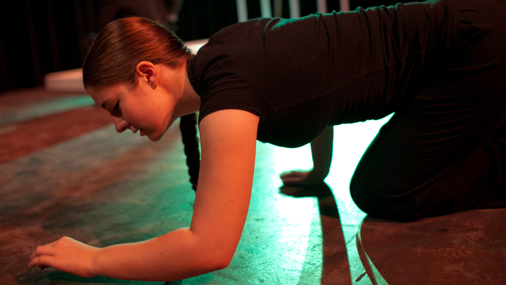 A girl wearing all black is crouched on her hands and knees as if she were crawling. She is looking at the floor. 