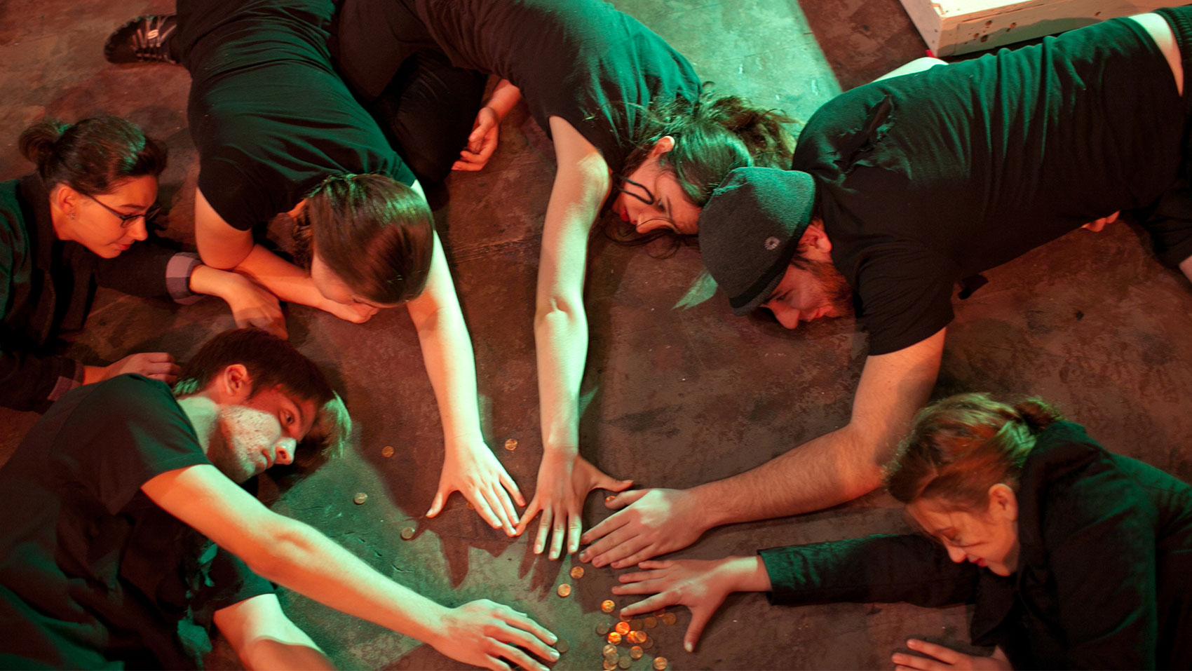 Six characters lay on the ground in a circle, all dressed in black, with arms outstretched toward coins scattered in the middle of the circle.