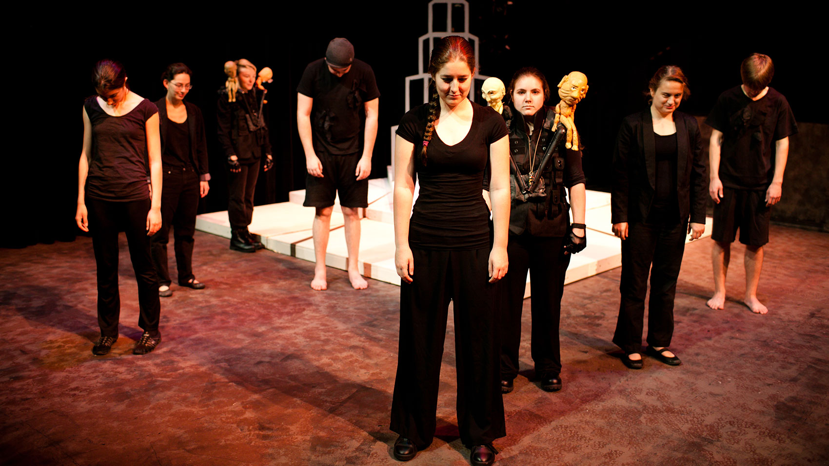 Eight characters stand in a clump with a tower structure in the background. The characters are all wearing black clothes, with two characters having a pair of dummies on their shoulders, and all of the characters are looking toward the floor. 