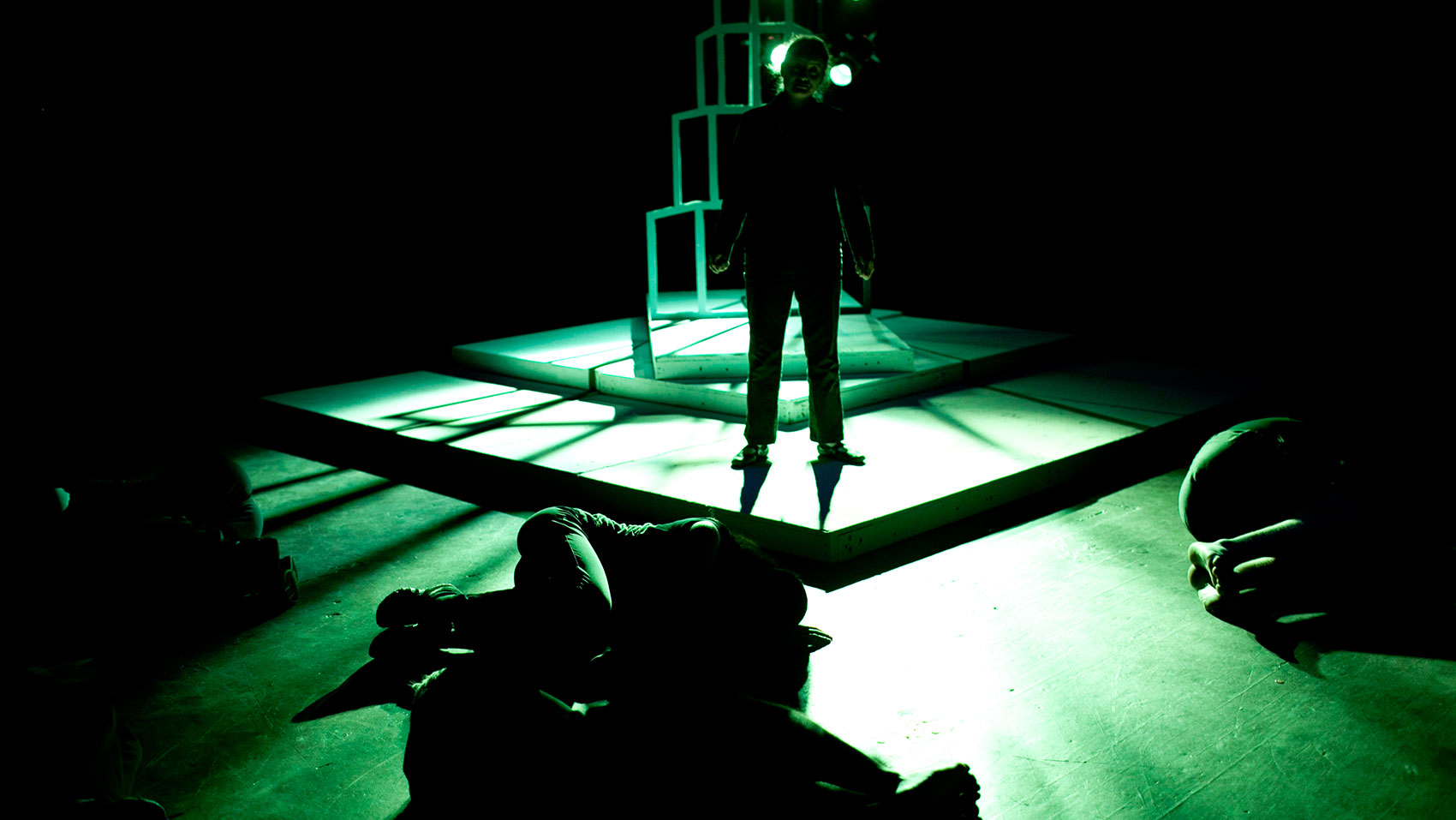 A character is silhouetted in green light in front of the tower structure, with other characters laying on the floor around her in fetal position.