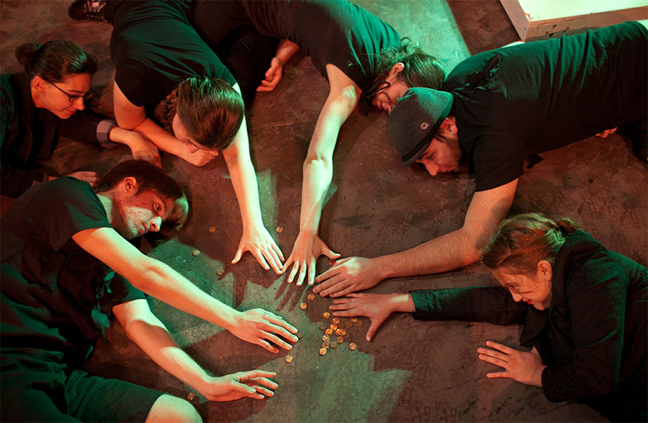 Six characters lay on the ground in a circle, all dressed in black, with arms outstretched toward coins scattered in the middle of the circle. 