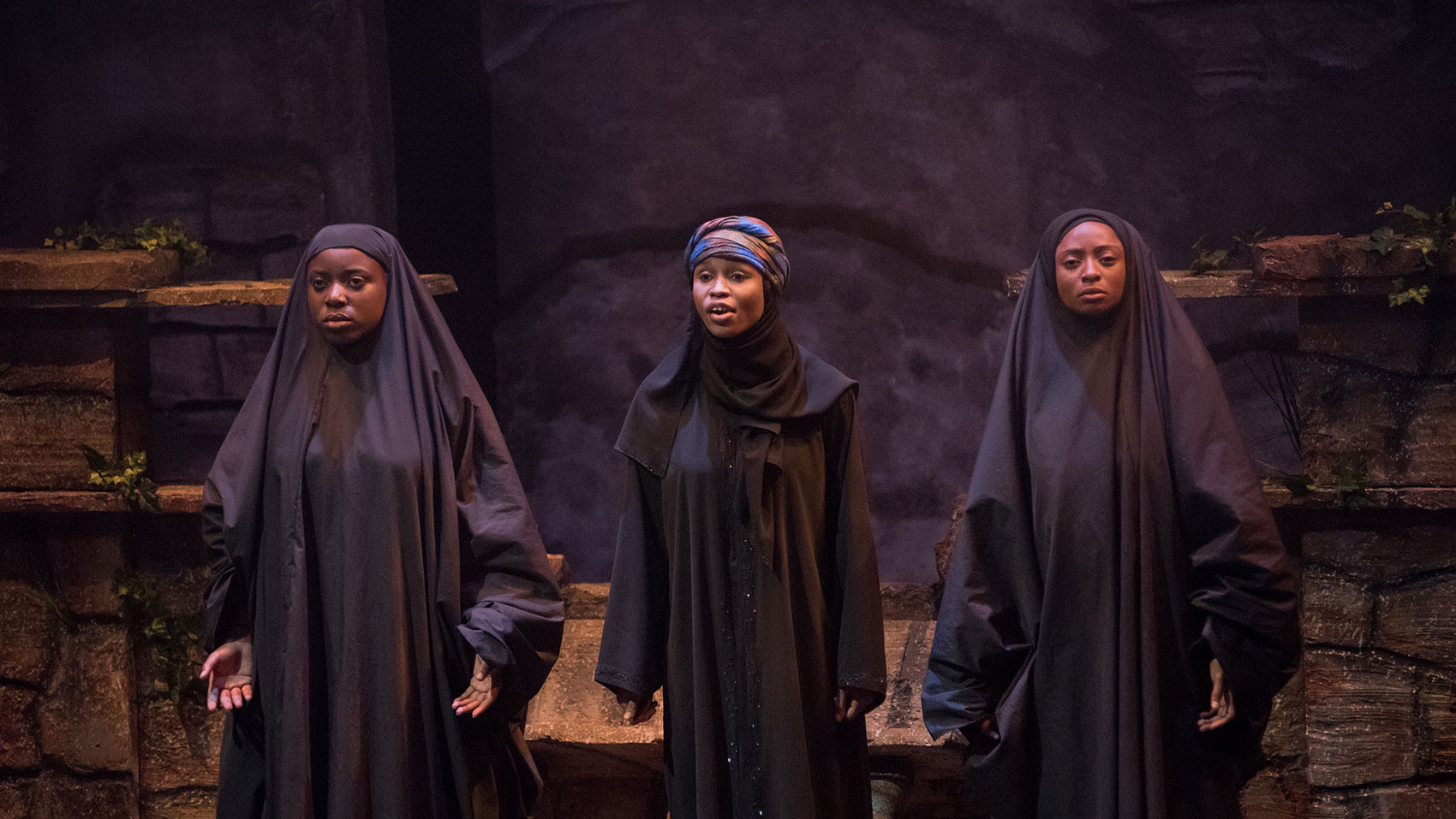 Three woman stand onstage in black, full-length body coverings, two with their hair covered by these coverings while one woman in the middle wears a colorful headscarf and is speaking. 