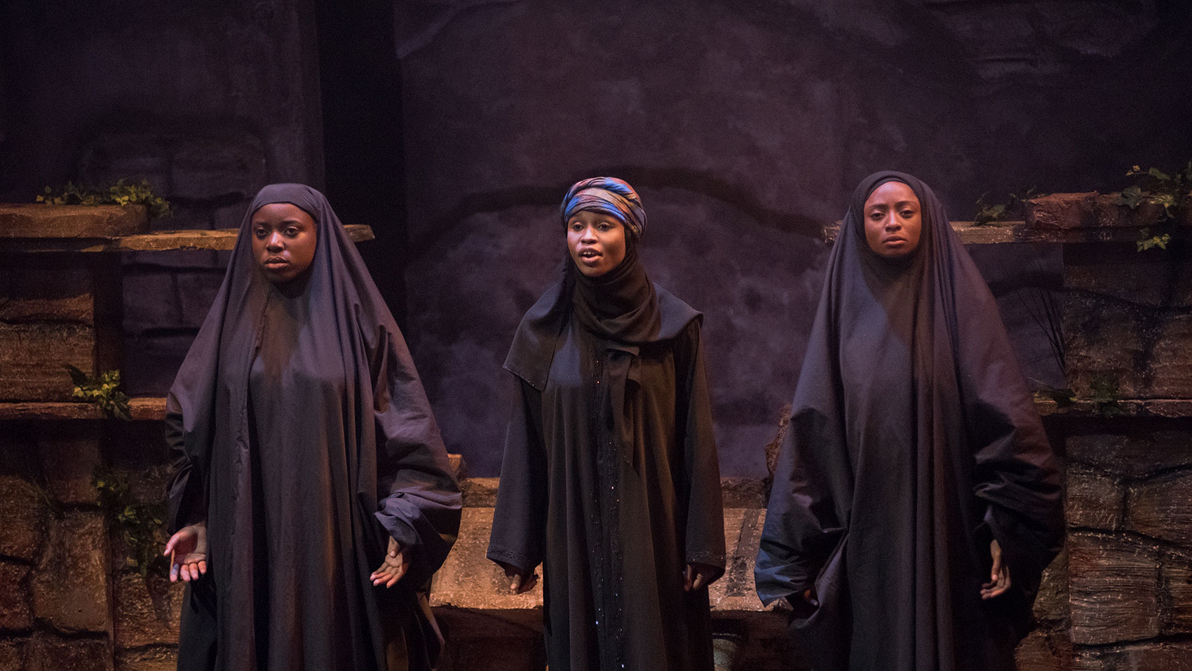 Three woman stand onstage in black, full-length body coverings, two with their hair covered by these coverings while one woman in the middle wears a colorful headscarf and is speaking.