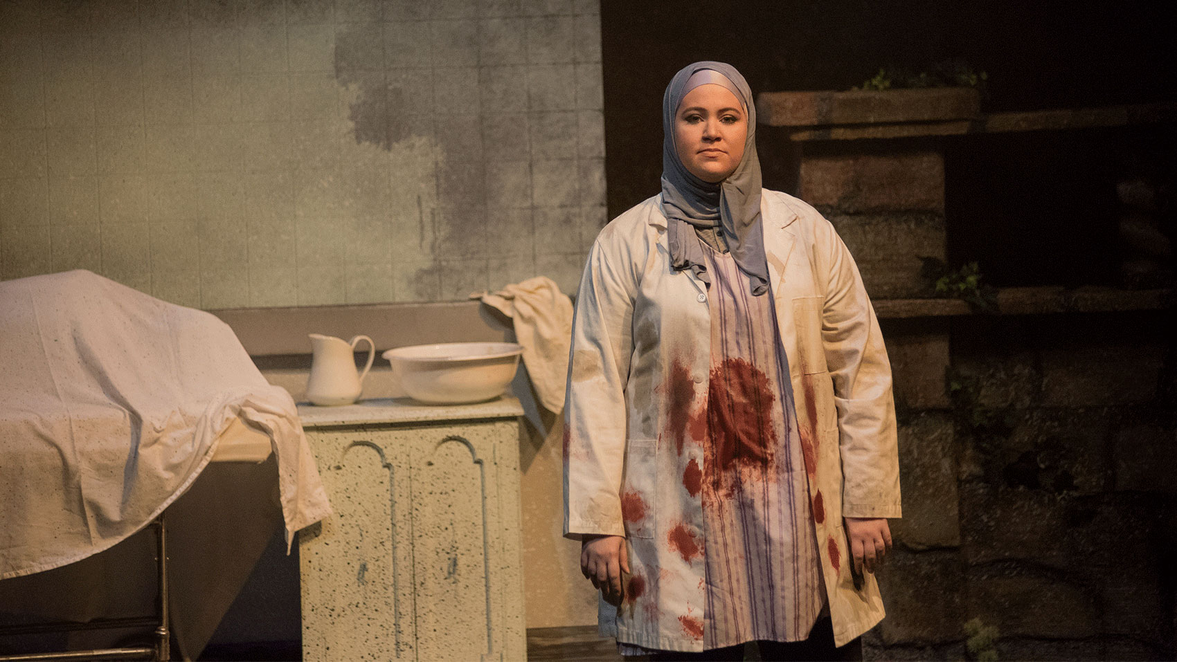 A woman in a hijab and white lab coat has blood soaked on her clothes, she has a somber expression on her face.
