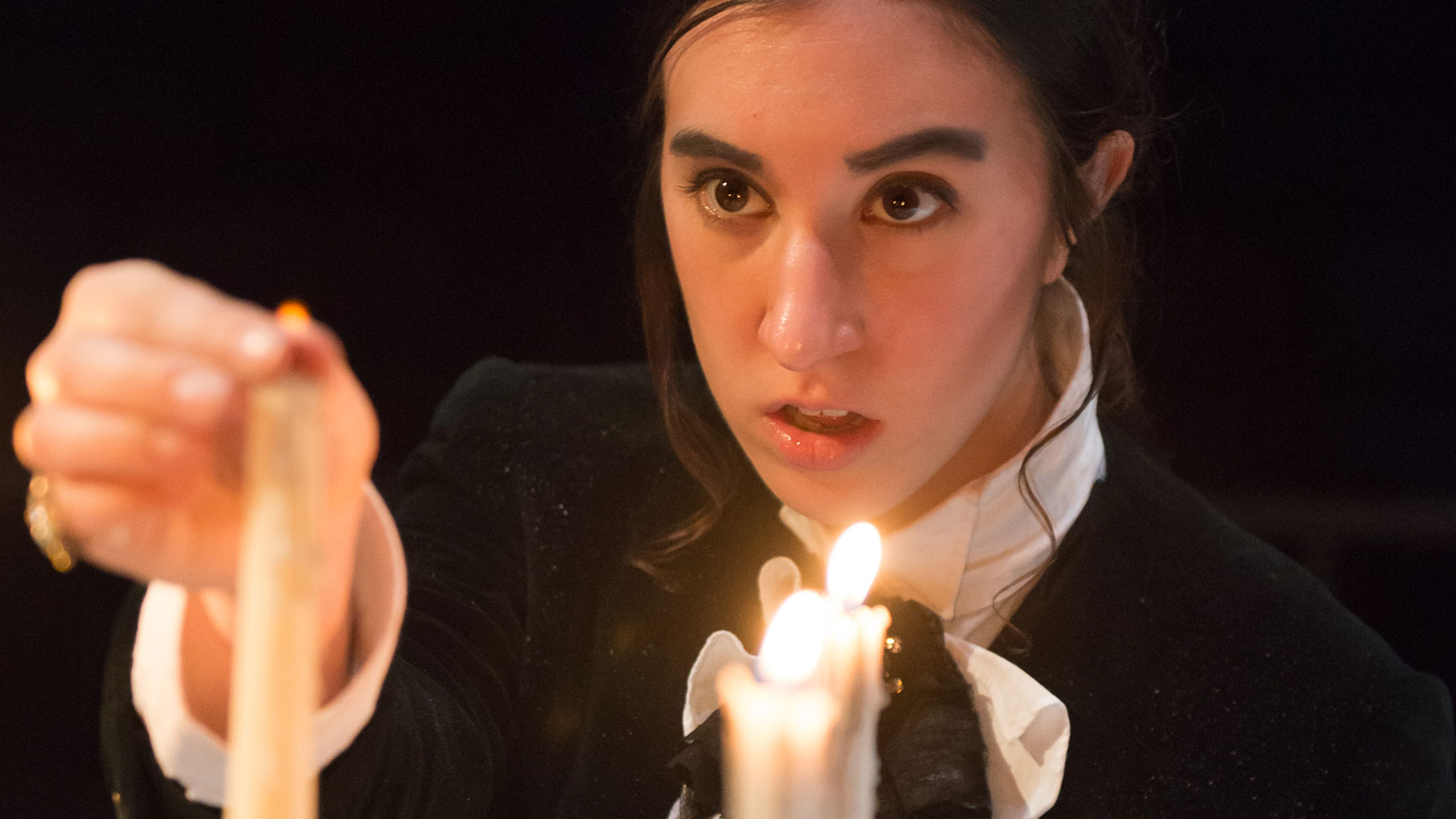 : A woman stands in front of candles, one is lit and the other is being snuffed out with her fingers.