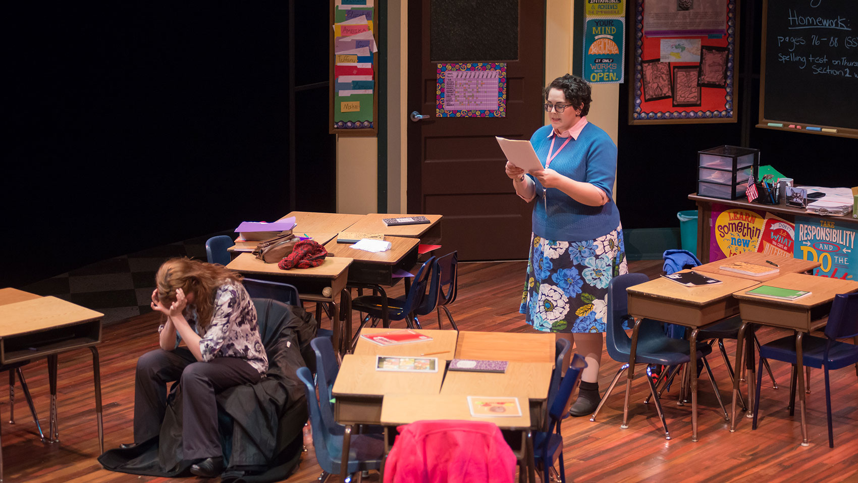 A teacher reads a document aloud to a woman, who is seated in a chair with her jacket underneath her as she has her face buried in the palms of her hands.