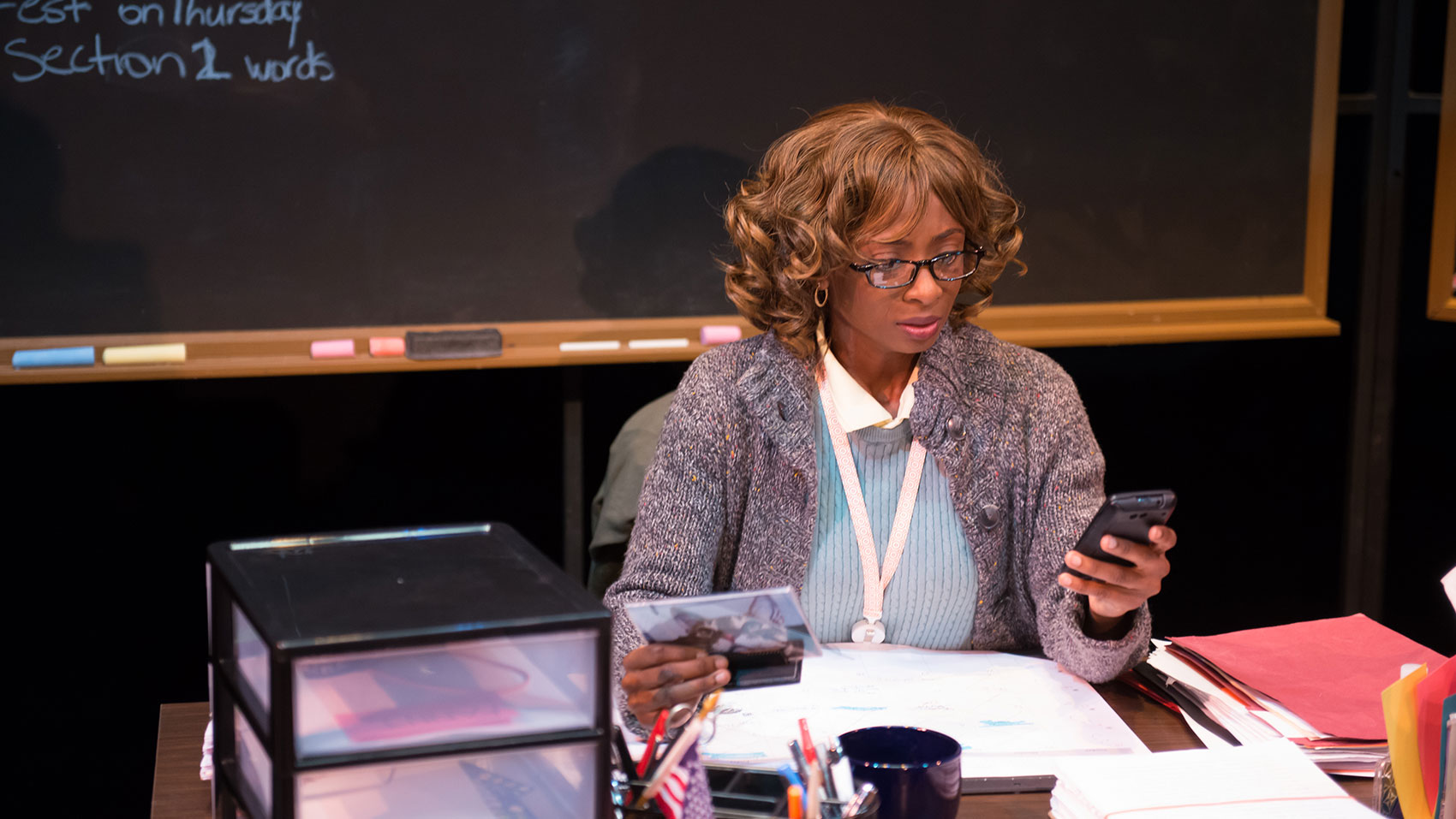 A teacher is seated at a desk, holding a CD case in one hand and is reading something on her phone, which is in her other hand.