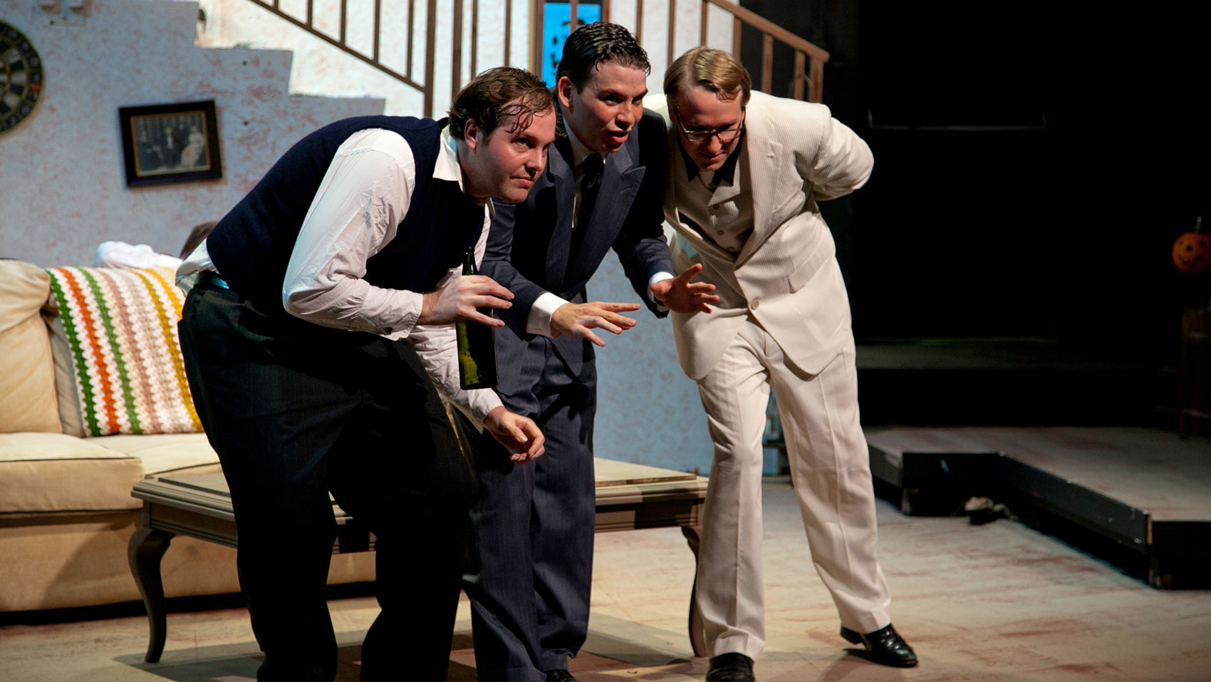 Three men are huddled near each other in conversation, one man has his hands raised low as if gesturing about something, the man on the left is leaning in toward the man in the middle to listen while raising a hand slightly while the man on the right is leaning in toward the man in the middle listening with a hand behind his back. 