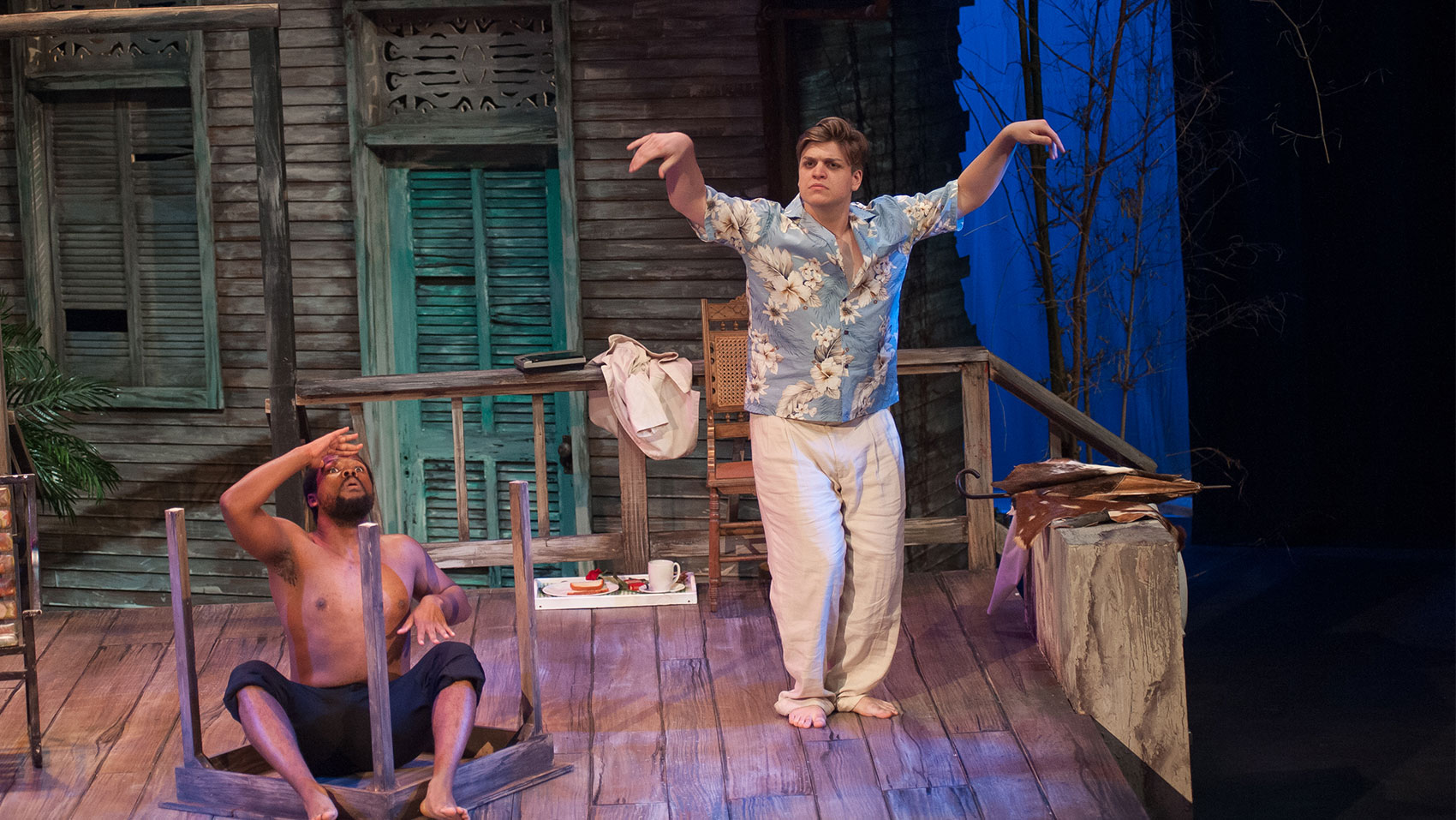 Two men are on a deck, one is sitting in an upturned table, leaning back with his hand over his forehead, eyes wide as if looking at something in the distance. The other man is standing, legs bent slightly and with arms raised and wrists hanging limply in the air.