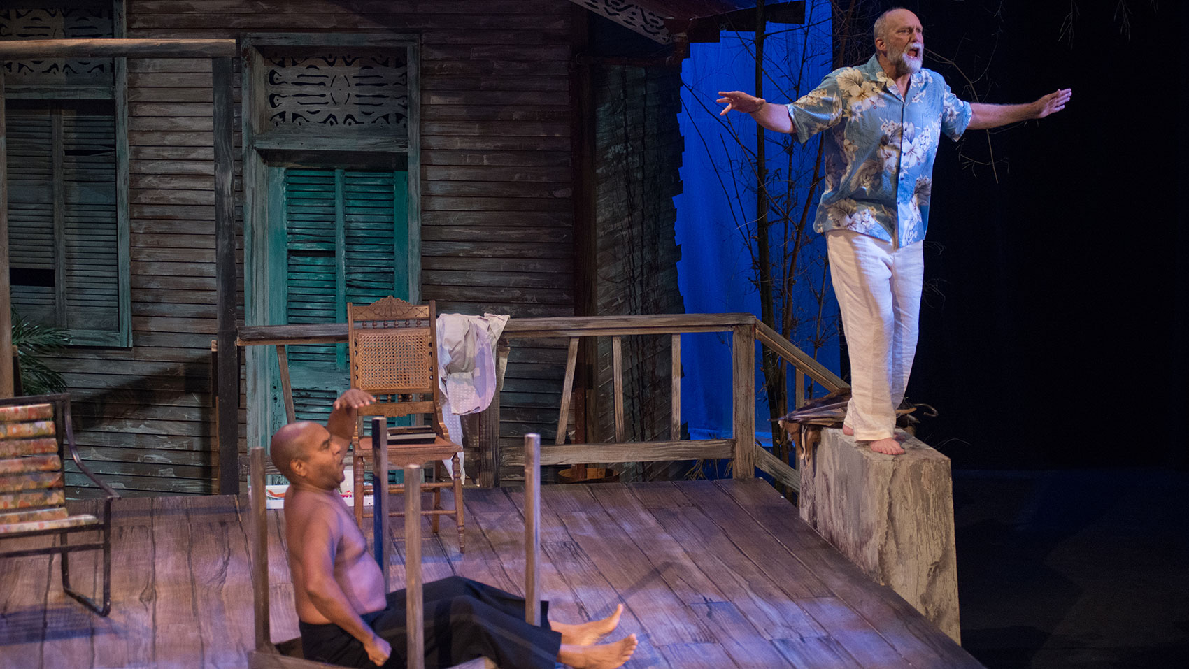 A man sits in an upturned table facing the right, hand in the air pointed horizontally as he is looking agape at something in the distance. Another man is standing on a railing of some kind, exclaiming and stretching out his arms as he keeps himself balanced.