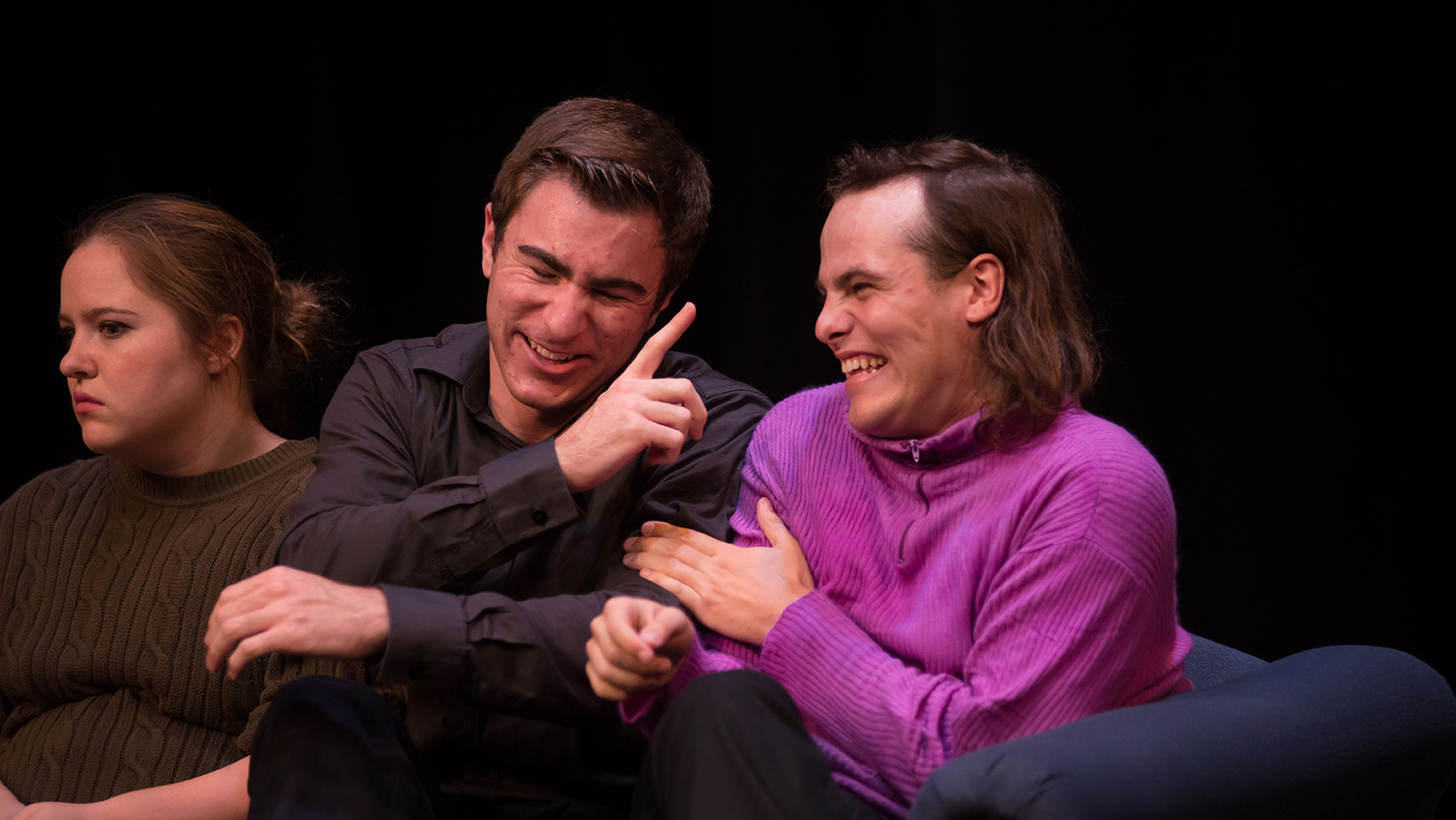 Three characters are on a couch. A woman stares off into space next to two men, the one in the middle of the three laughing and gesturing his finger while the man on the right laughs and has one hand on the first man’s arm.
