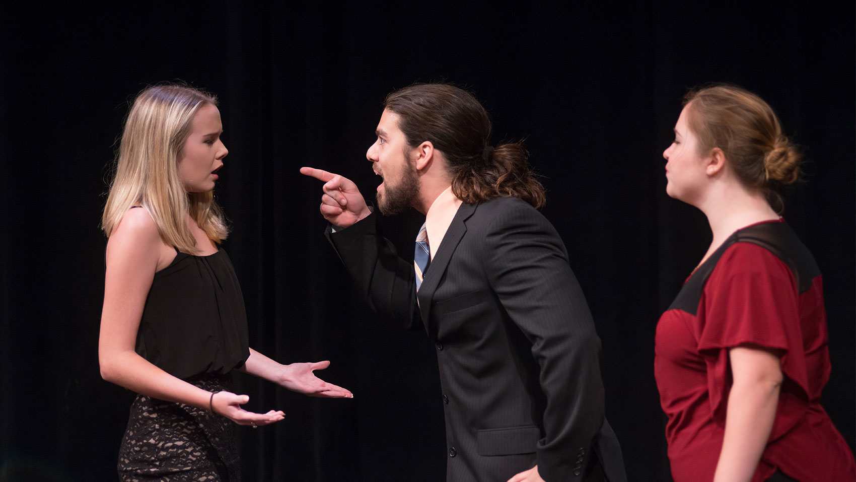 A man is yelling and pointing a finger in accusation at a woman, who looks upset and has her palms facing upward in confusion. Another woman stands behind the man watching the exchange.  