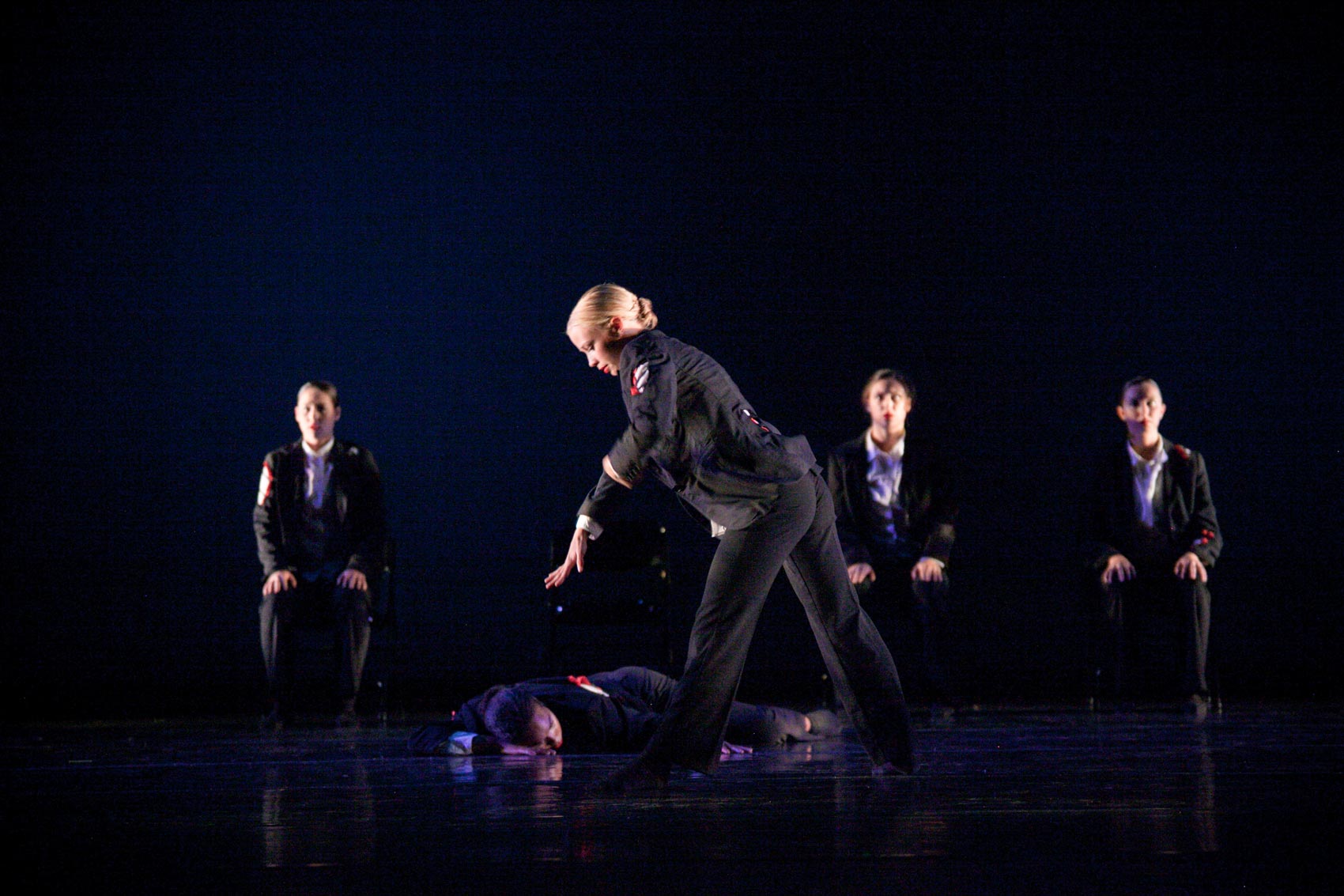 A dancer reaches down to another dancer on the floor while several others sit in the background.