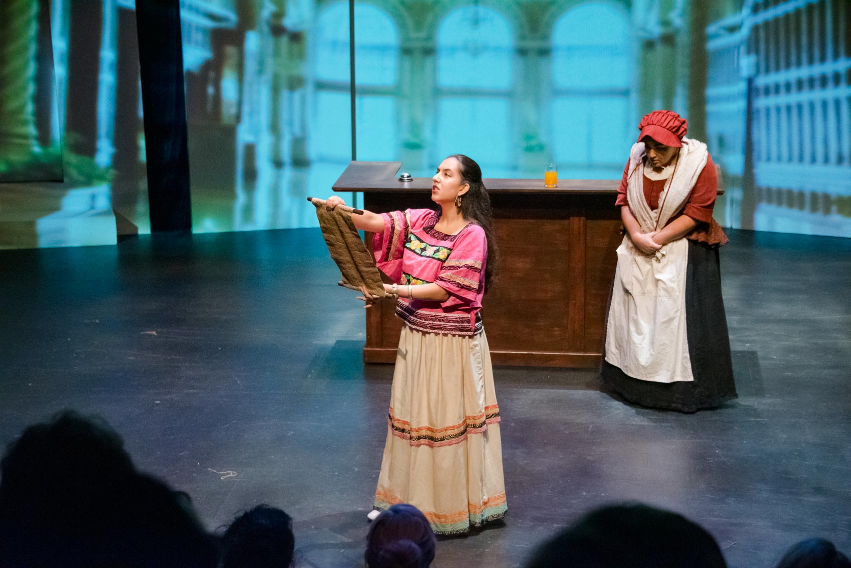 A woman reads from a scroll in front of a servant.