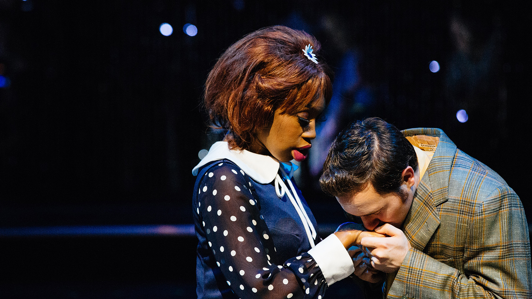 A Black woman wearing a navy dress with a white collar and white polka dots on the sheer sleeves gets her hand kissed tenderly by a man in a plaid jacket. He holds her hand with both his hands. She looks at him kissing her hand.