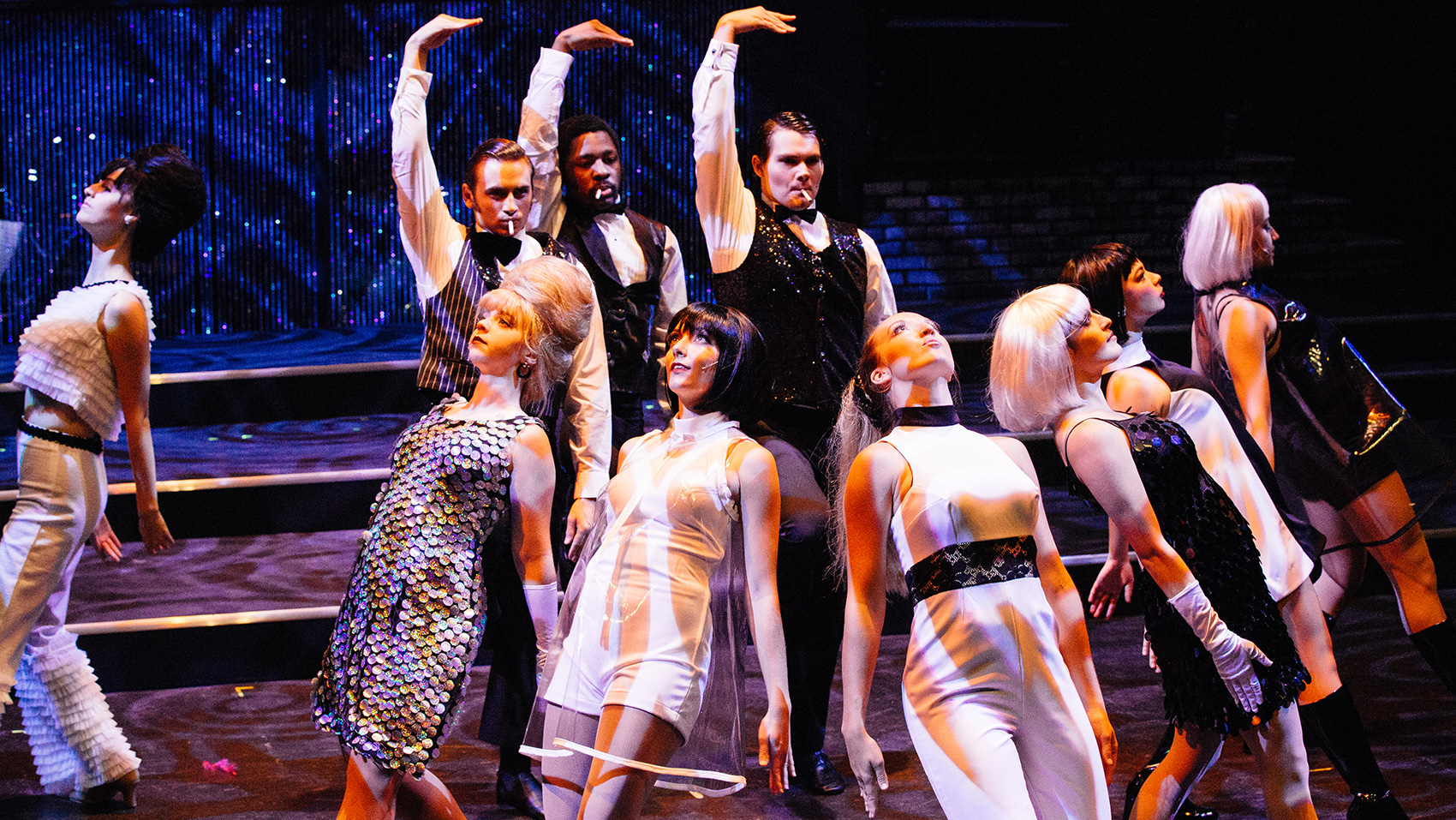 Three men wearing white dress shirts, black dress pants, bow ties and vests raise their right arm above their head, their hands bent to be parallel to the ground. Seven female performers, all in dressy retro attire, bend their bodies slightly backwards so that they look up to the ceiling, they keep their arms straight to their sides.
