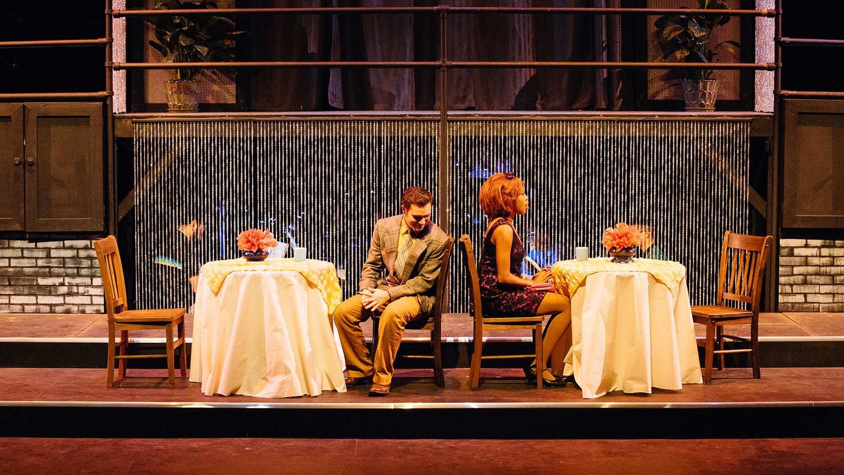 There are two small round dining tables with two chairs each. It is a dressy restaurant, white tableclothes and a small vase of flowers on the table. A man and and a Black woman are seated separately, the latter sits with her back facing the man, as he turns slightly so that he is facing the side. It looks as if he is talking to her.