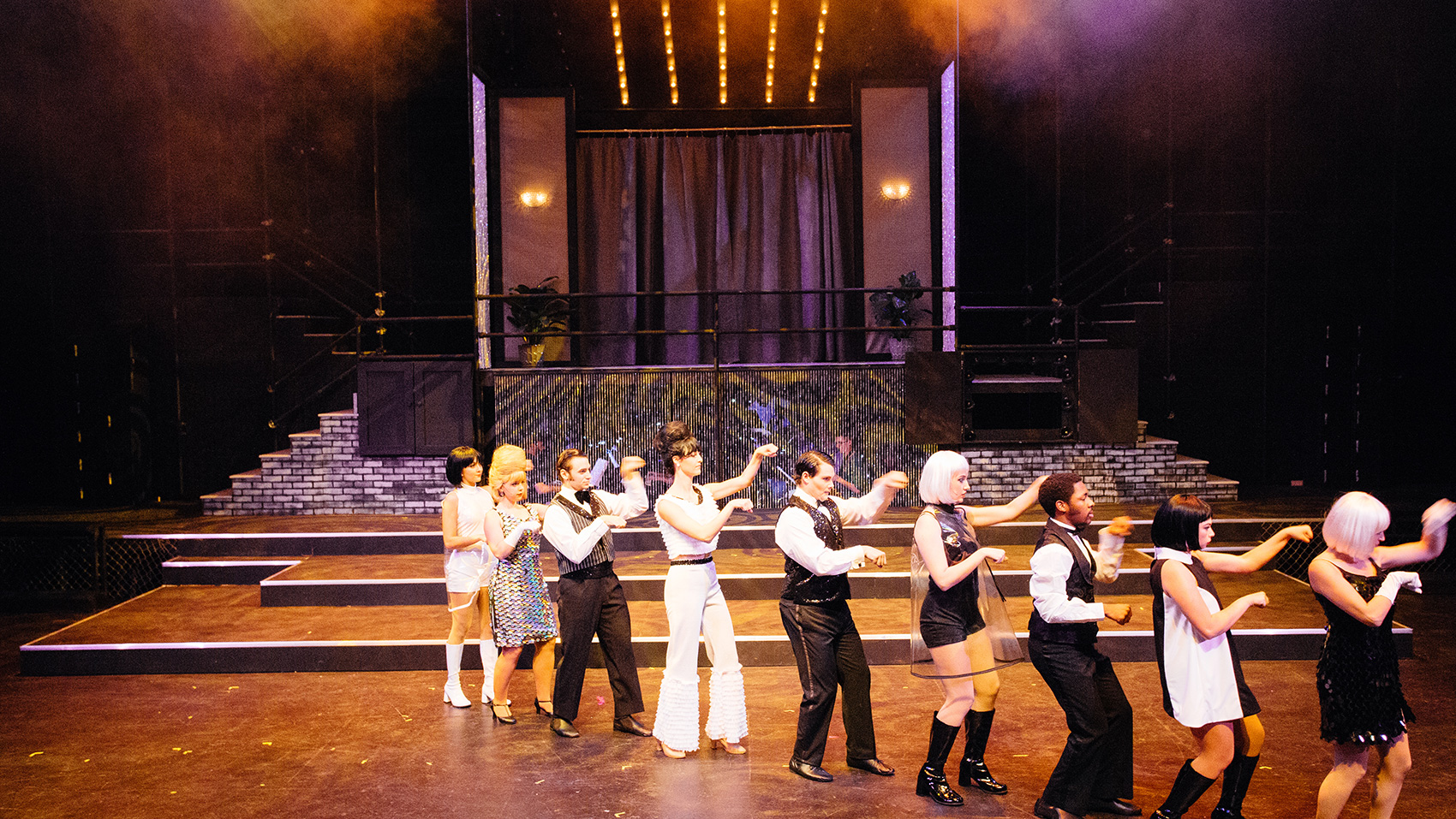 A group of performers, male and female, dance in a single line. Behind them stands a raised platform designed to look like the entrance to a fancy and glitzy retro sott of hall or restaurant, with purple curtains, soft lights, and a short flight of stairs on either side of the platform.