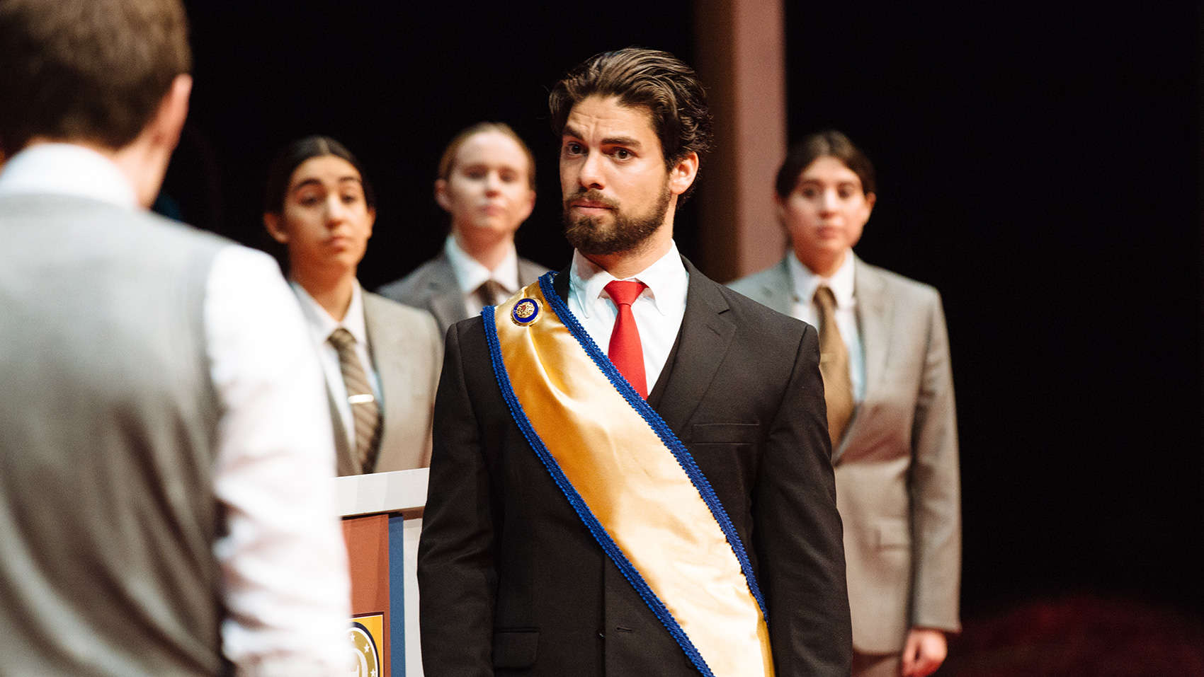 A man wearing a red tie and a yellow-gold sash with blue trim stands facing another man, whose back is facing us. The man in the rd tie has a quizzical eyebrow raised. Three women wearing suits and ties stand behind them, watching.