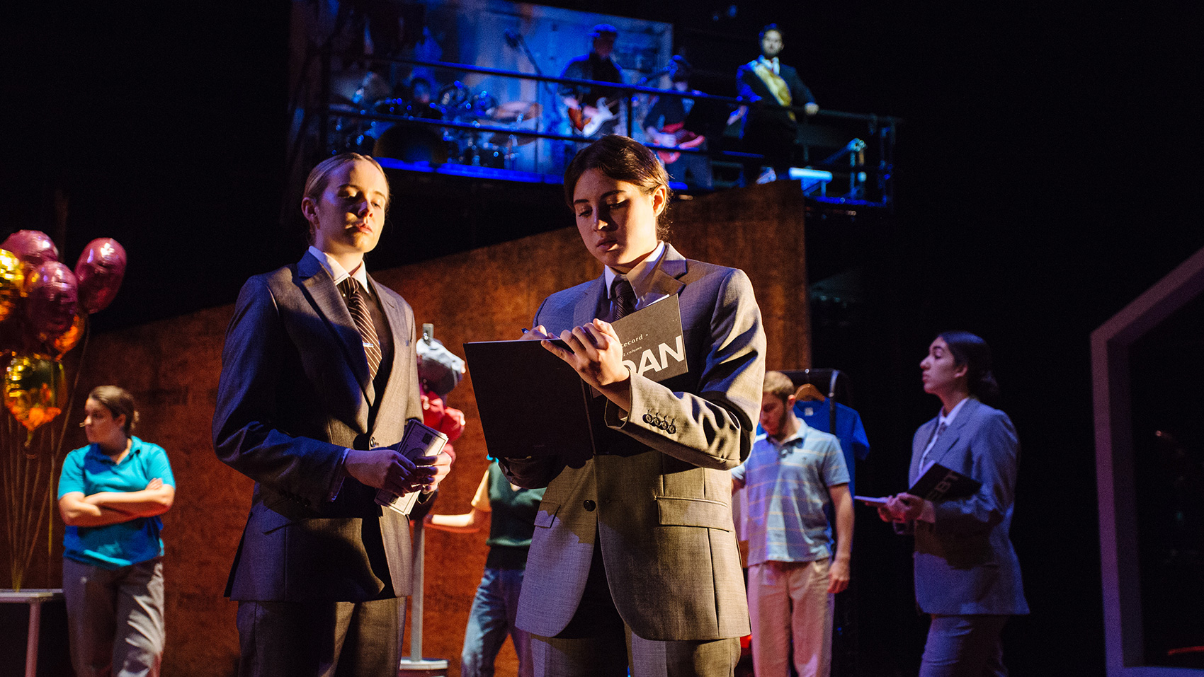 Two women in suits and ties confer together, one of them writing notes in a book. 