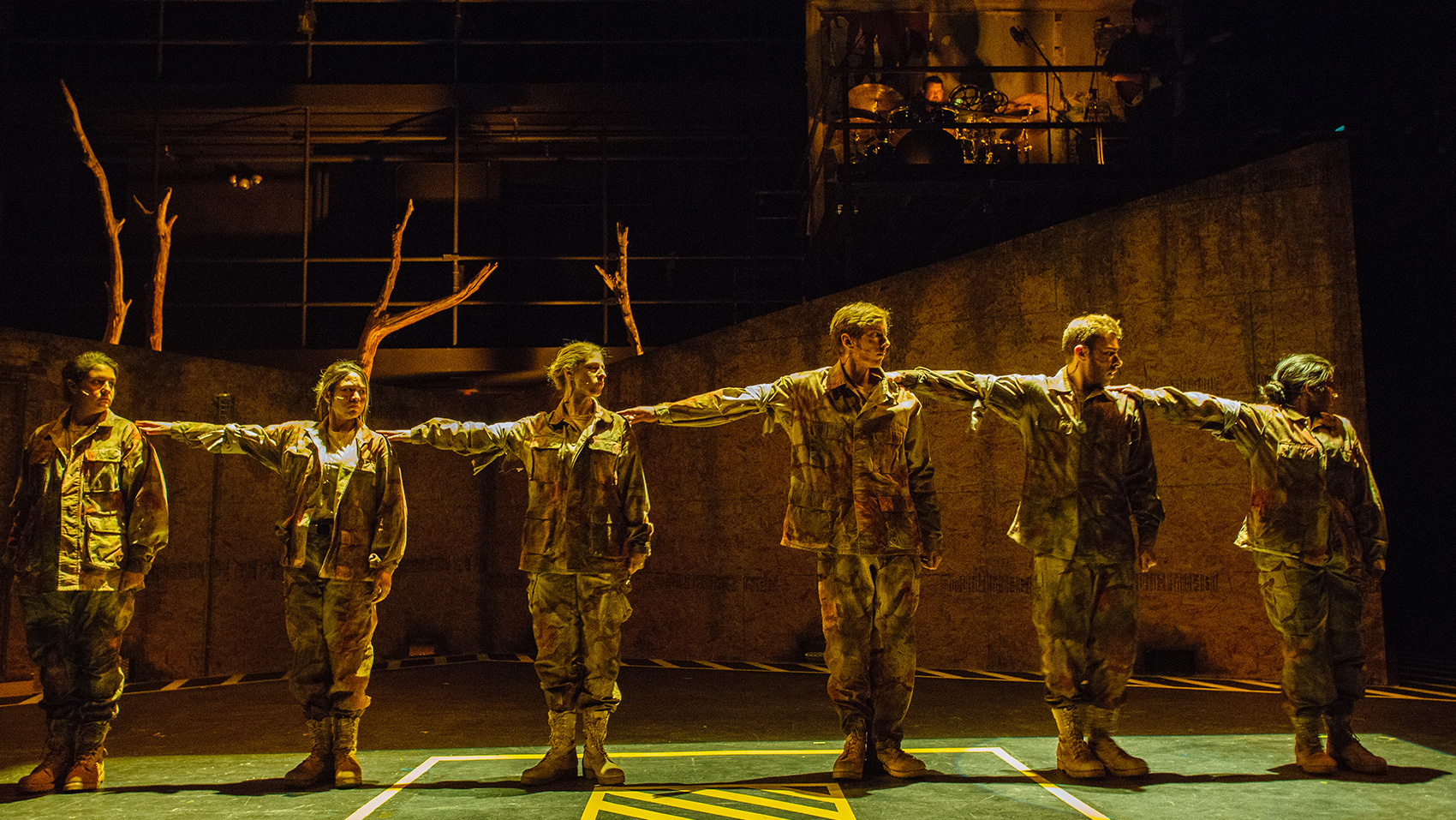 Six people stand in a straight line, each touching the one behind them on the shoulder with their arm stretched straight to the side. They are all in camo wear.