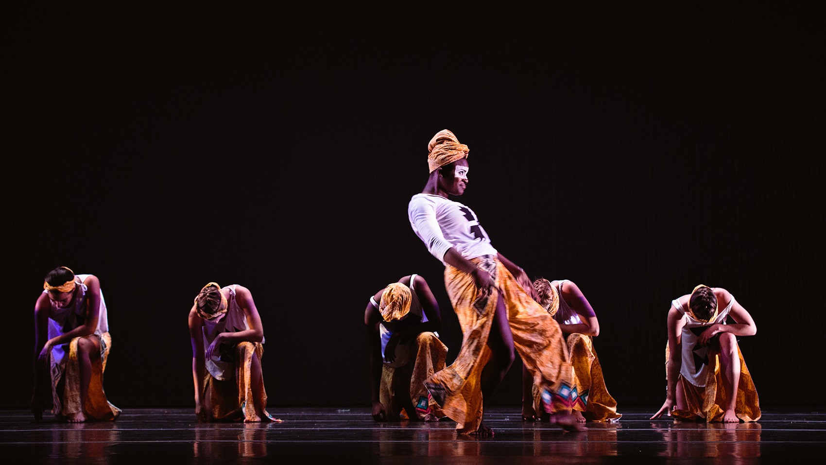 Five female dancers kneel on one knee, with one hand they touch the ground as they rest their other arm on leg that's not kneeling. They all bow their hands. In the foreground a solo dancer dances. All dancers are wearing traditional African prints.