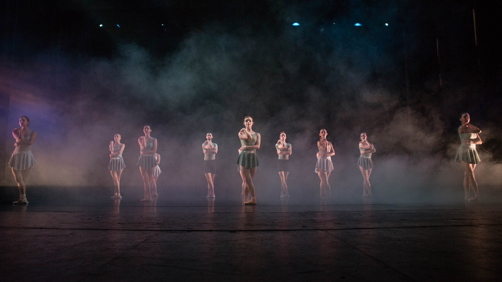 Female ballet dancers hold the same pose onstage, a haze of atmospheric lighting and smoke flowing over them.